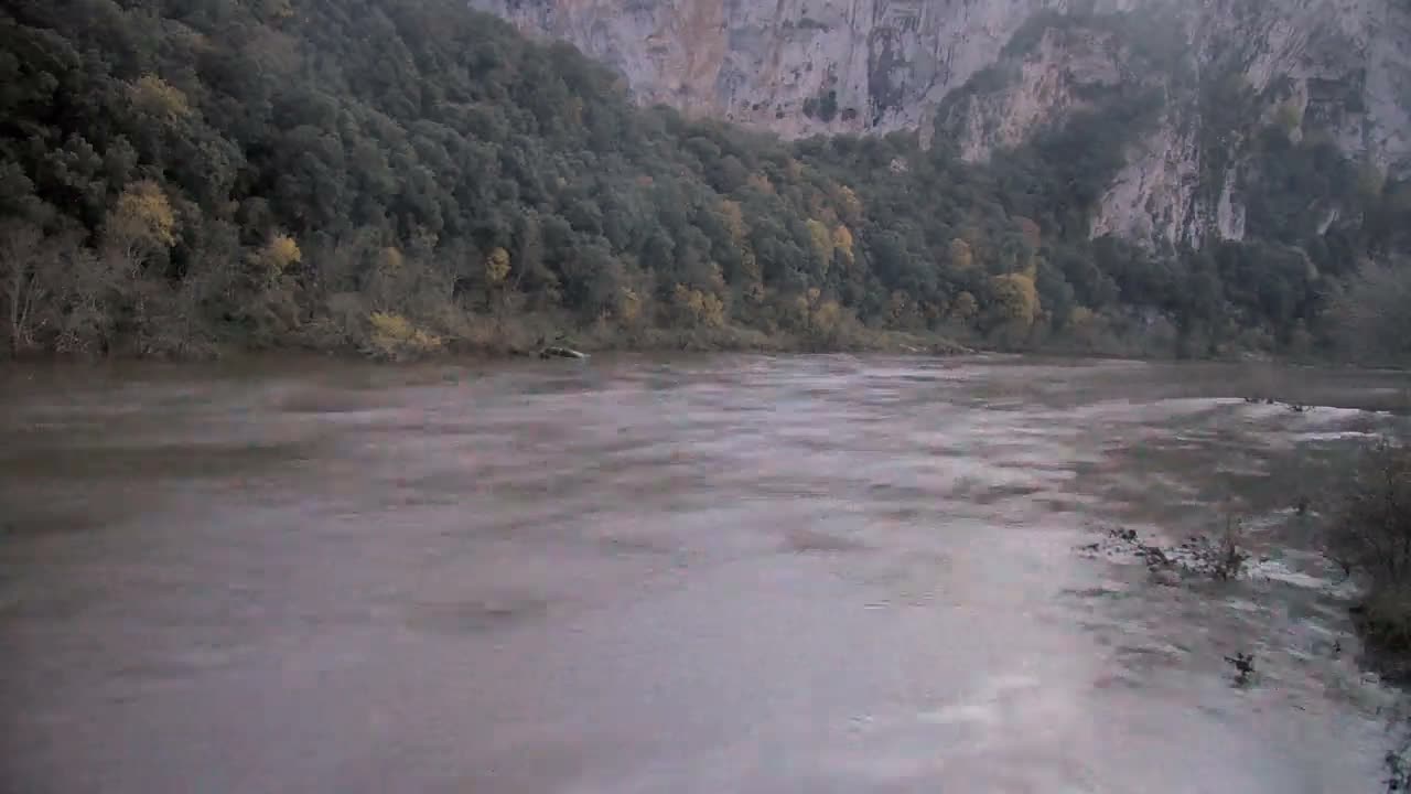 A calm river flows through a rocky, wooded valley under a sunny sky, with several small boats visible on the water.