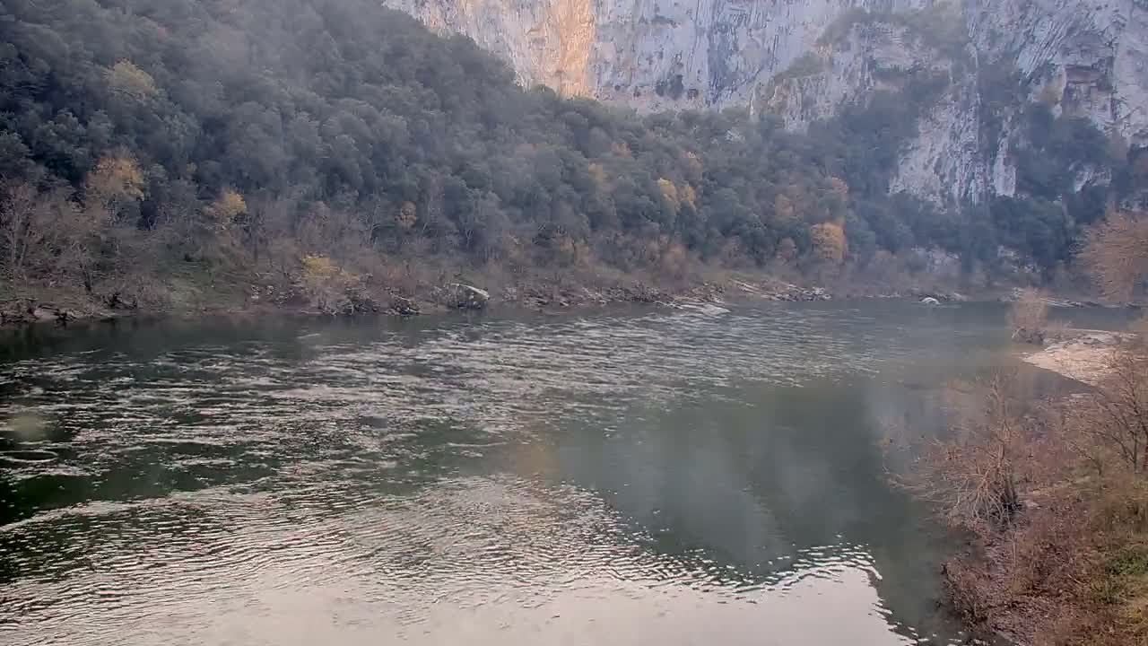 A wide, rippling river flows through a valley flanked by a densely forested hillside on one side and towering rocky cliffs on the other, all under an overcast sky.