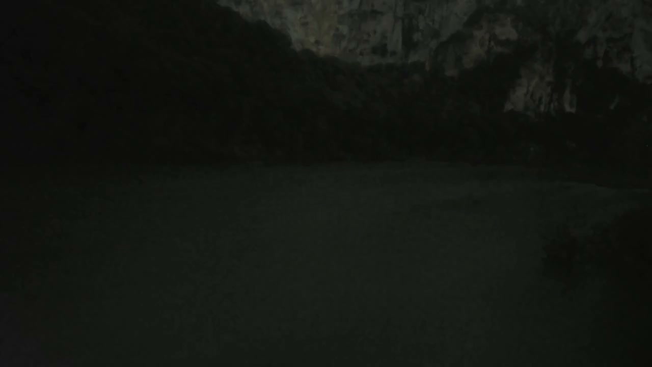 A wide, rippling river flows through a valley flanked by a densely forested hillside on one side and towering rocky cliffs on the other, all under an overcast sky.