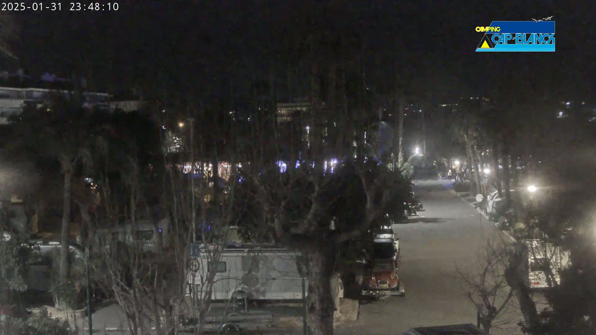 A nighttime, dimly lit road winds through a campground, lined with parked vehicles and trees.