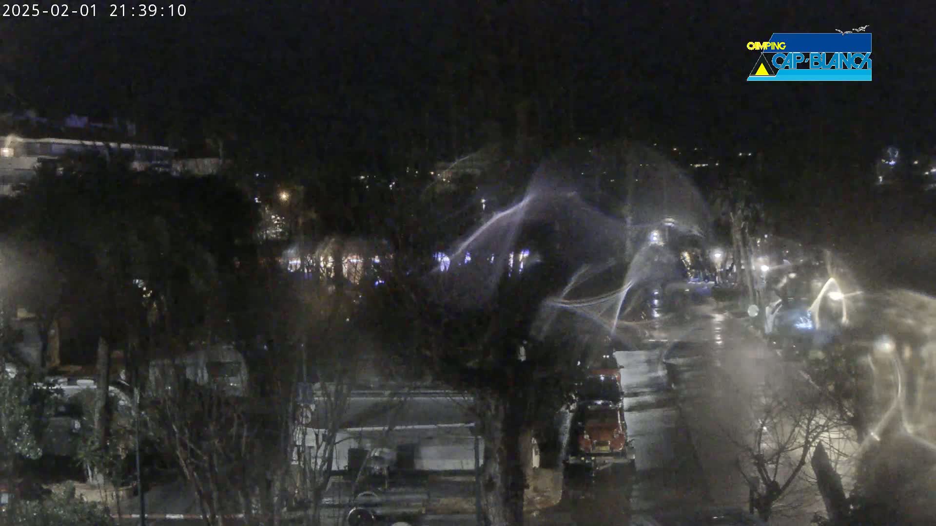 A nighttime, rain-swept view of a campground with vehicles and trees, illuminated by scattered lights.
