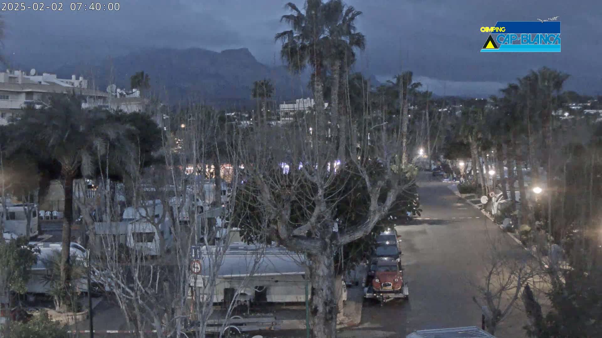 A street lined with palm trees and recreational vehicles is visible under a dark, cloudy sky with mountains in the distance.