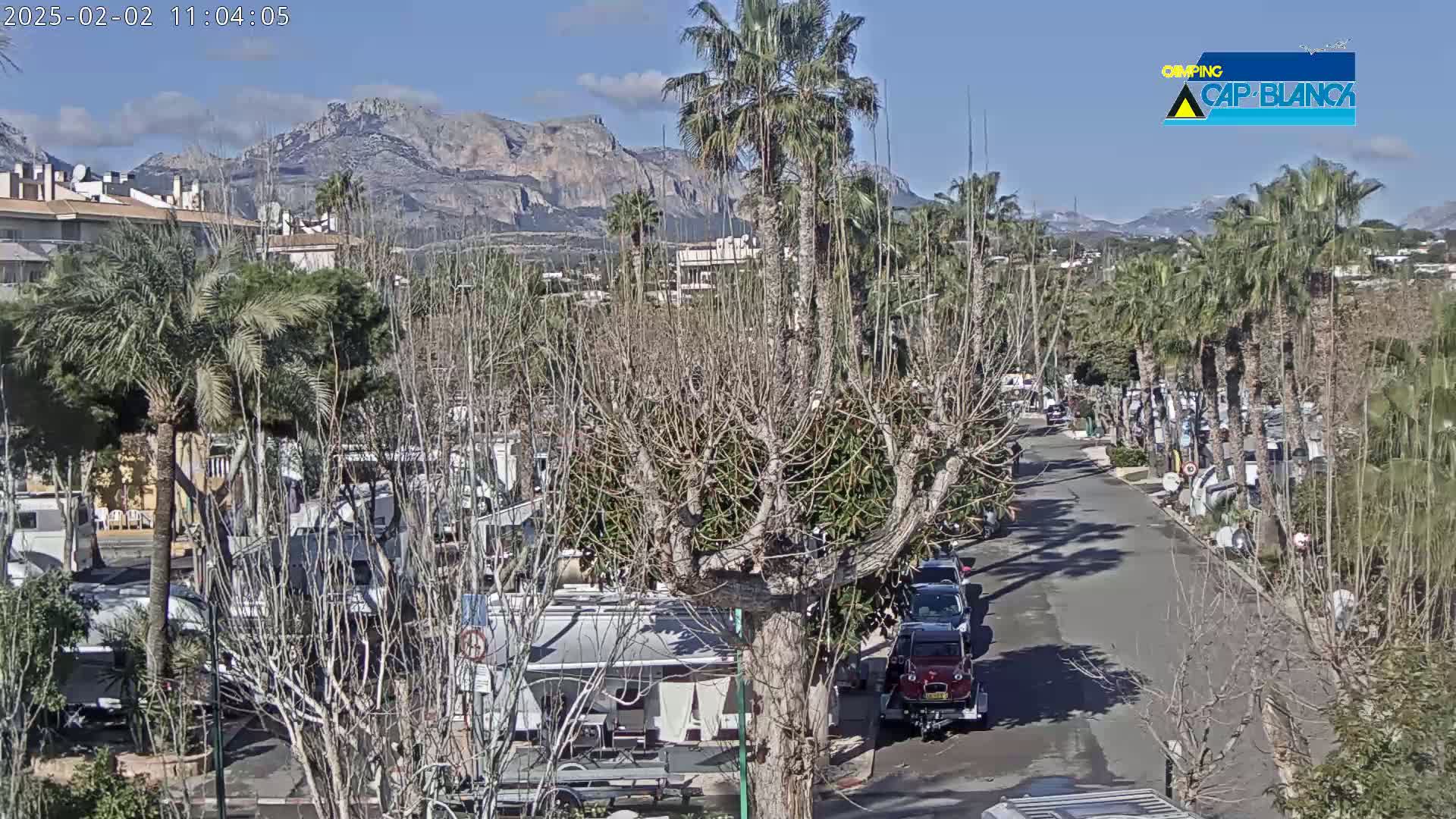 A sunny day reveals a paved road lined with parked RVs and palm trees, leading towards a mountain range in the background.