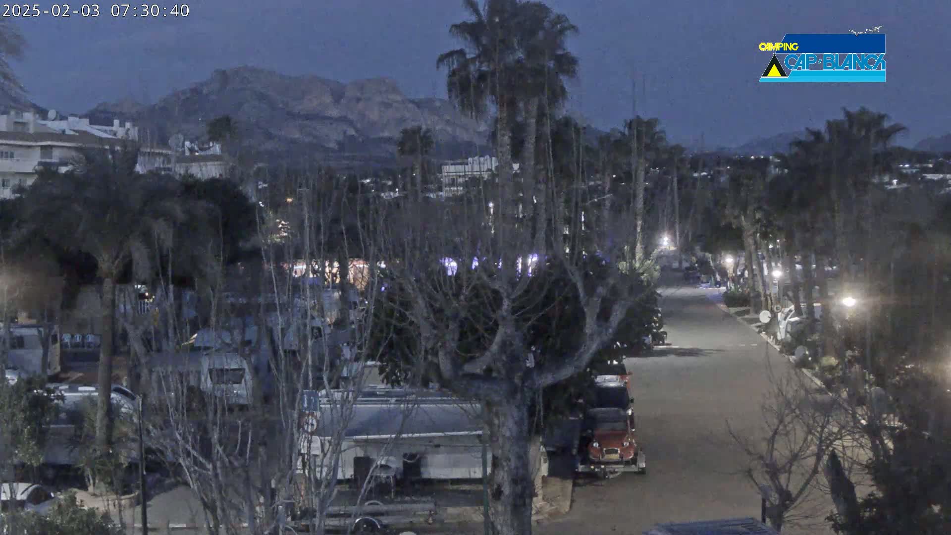 A nighttime view of a campground with RVs parked along a paved road, under a twilight sky, with mountains in the distance.
