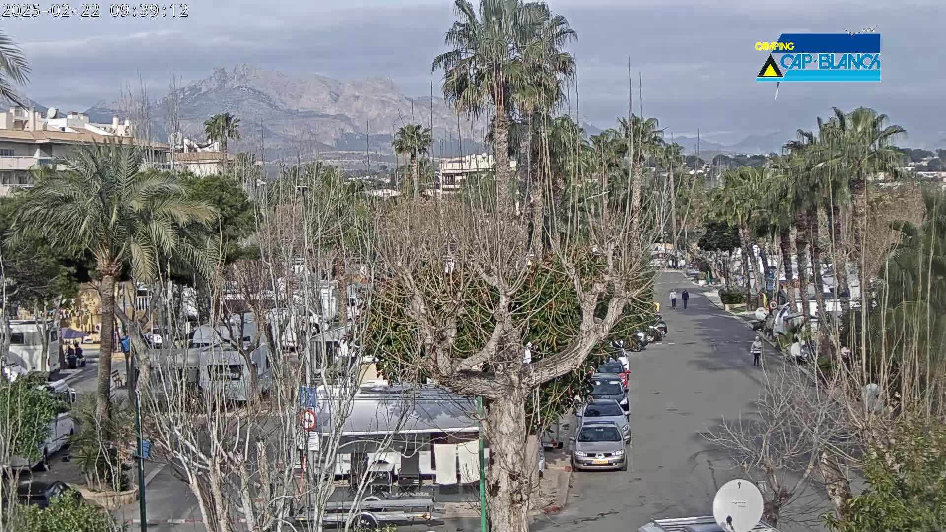 A mostly sunny day reveals a street scene with parked RVs and cars, palm trees, and leafless trees, set against a backdrop of mountains.