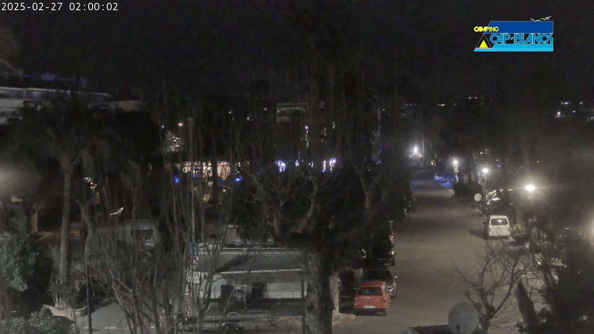 A nighttime view of a street with parked cars and RVs, surrounded by trees and buildings under dark, clear skies.