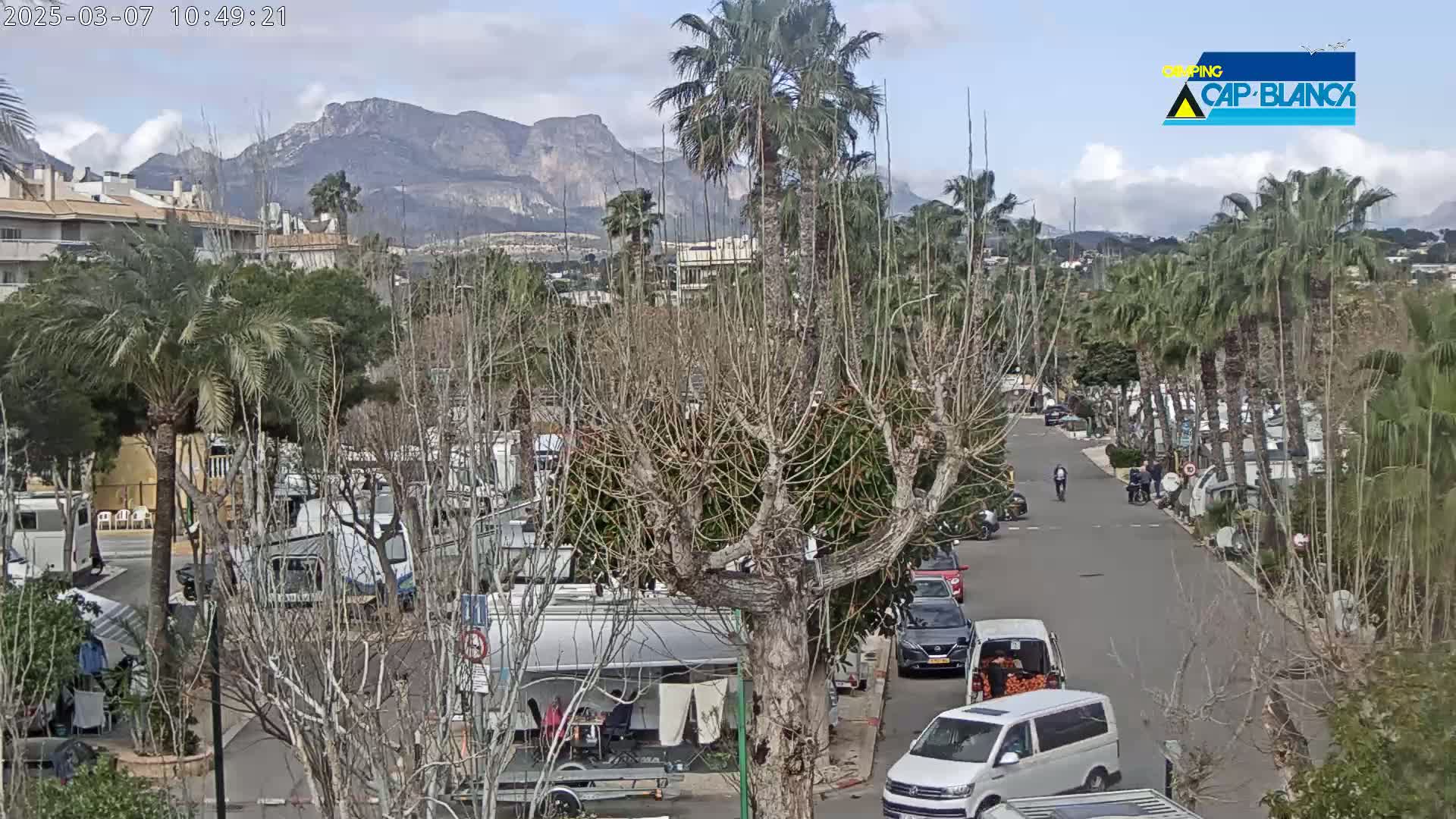A mostly sunny day shows a street scene with RVs, palm trees, and barren deciduous trees in front of mountains.