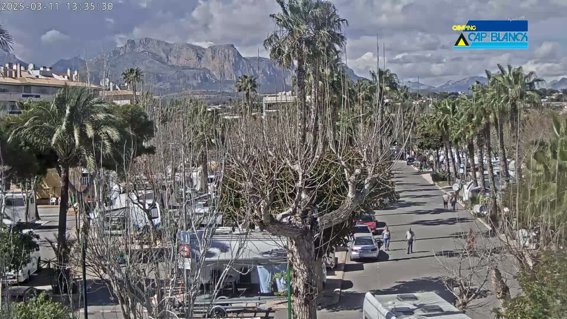 A partly cloudy day reveals a street scene with palm trees, parked RVs, and cars, all set against a backdrop of mountains.