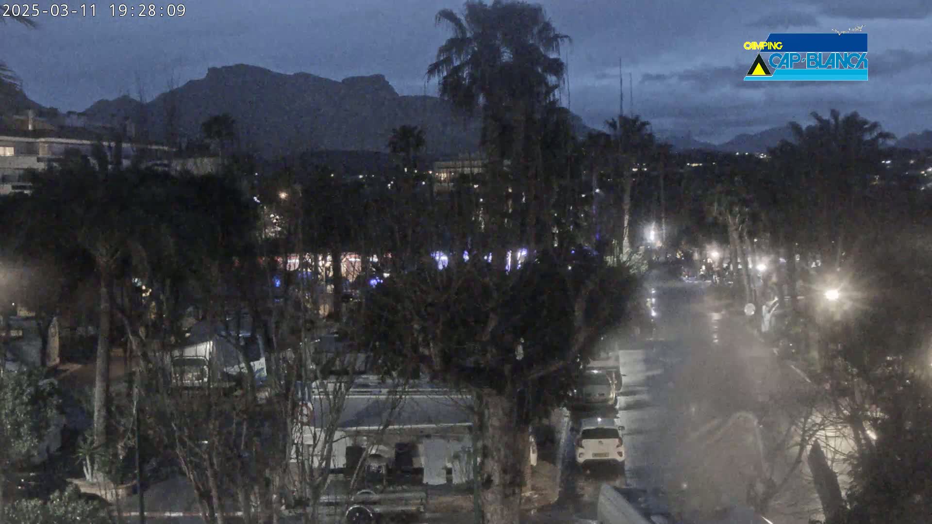 A nighttime view of a residential area with palm trees and mountains in the background, under a dark, cloudy sky.