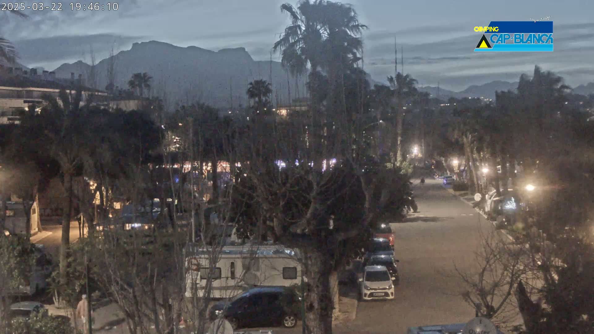 A nighttime view of a street lined with parked cars and recreational vehicles, under a dusky sky with mountains in the background.