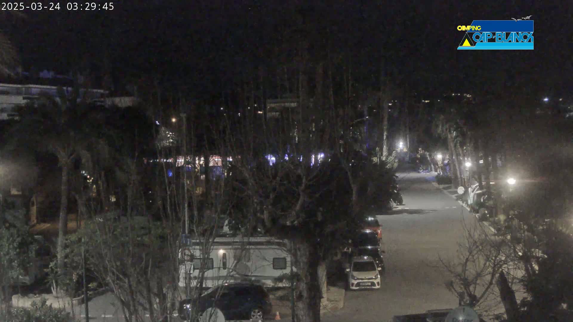 A nighttime view of a paved road lined with parked cars and recreational vehicles, surrounded by trees and buildings under a dark sky.