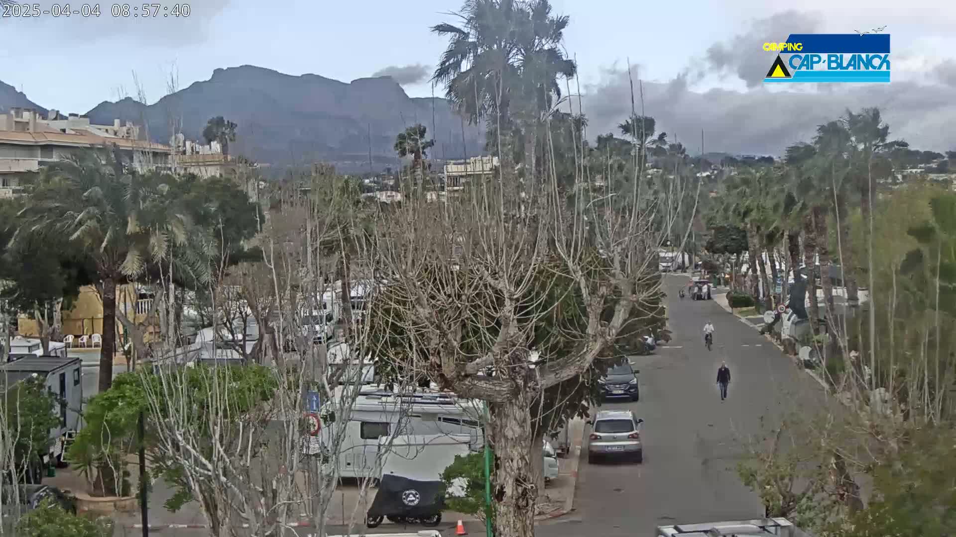 A street scene shows a cloudy day with several RVs parked among bare trees and palm trees, with mountains in the background and a few people walking and biking on the street.