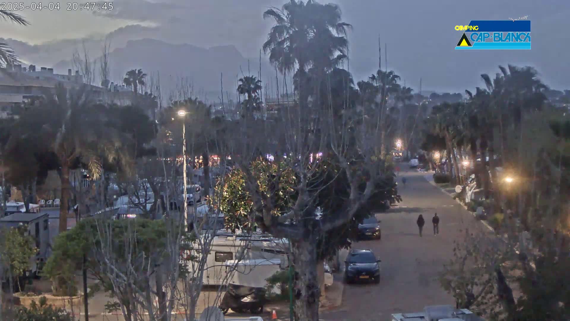 A nighttime view of a road lined with parked cars and RVs, under a dusky sky with mountains in the distance, shows two people walking along the road.