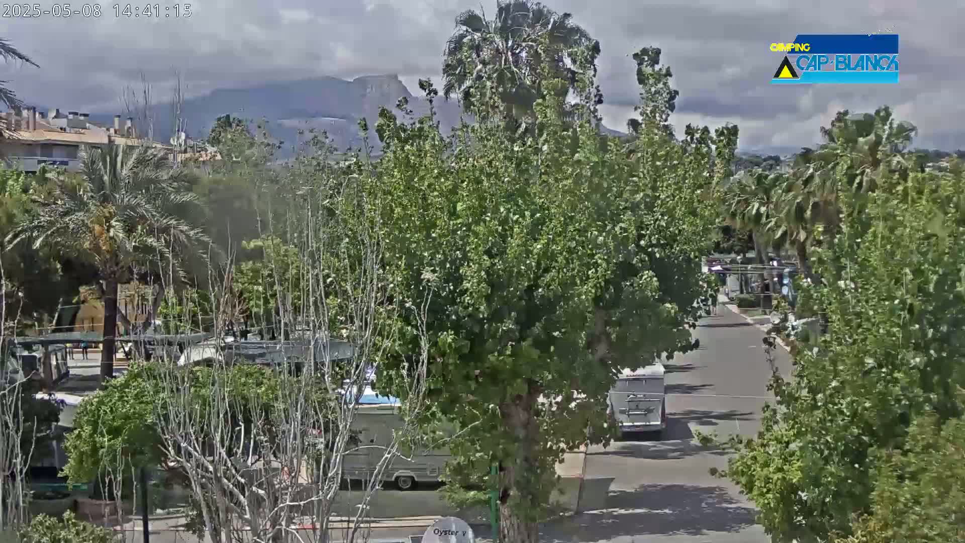 A partly cloudy day reveals a street scene with palm trees, buildings, and camper vans, mountains visible in the distance.