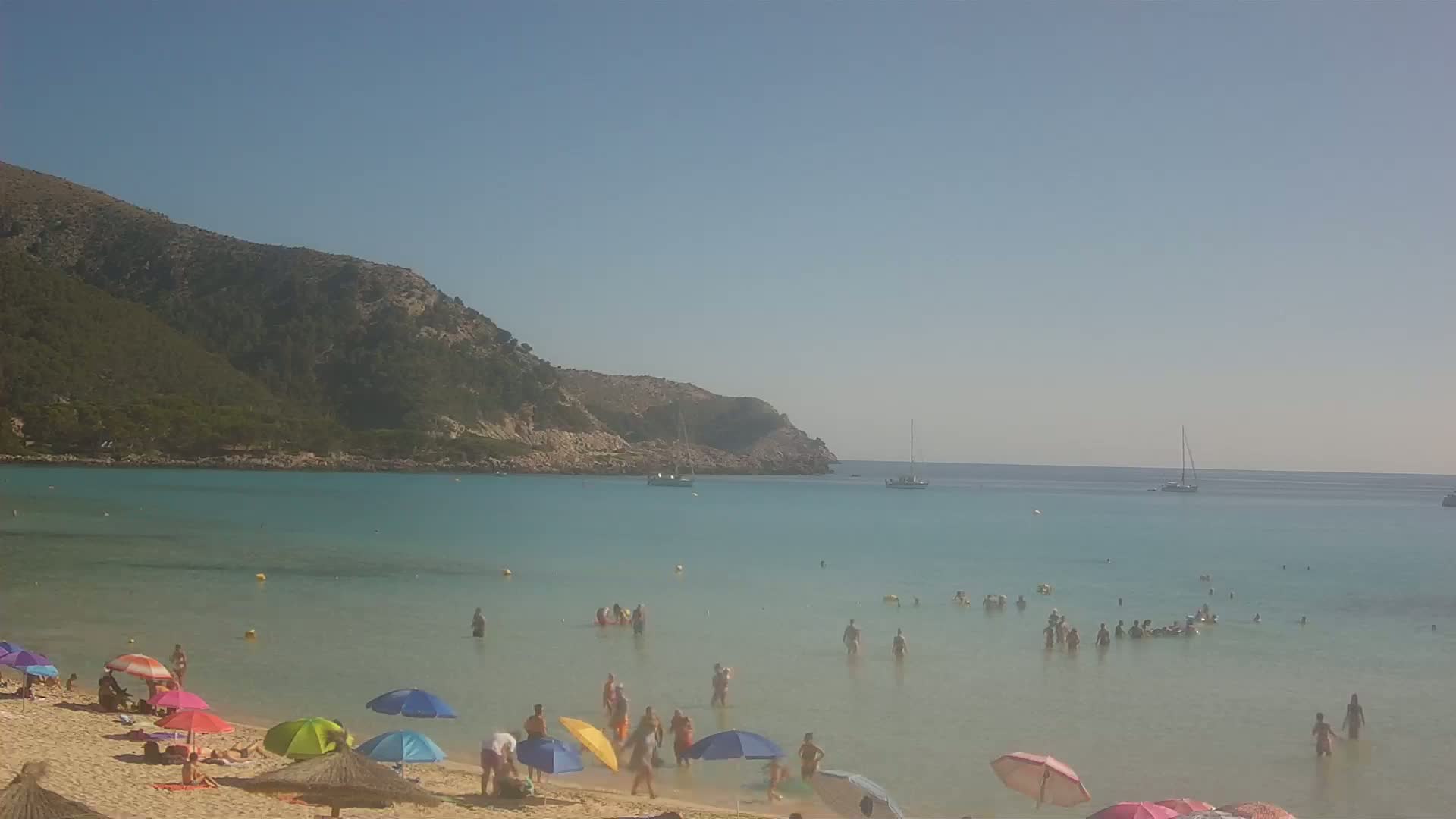 A sandy beach with colorful umbrellas and many people swimming in clear, shallow turquoise water, backed by a rocky, green hill under a clear blue sky.