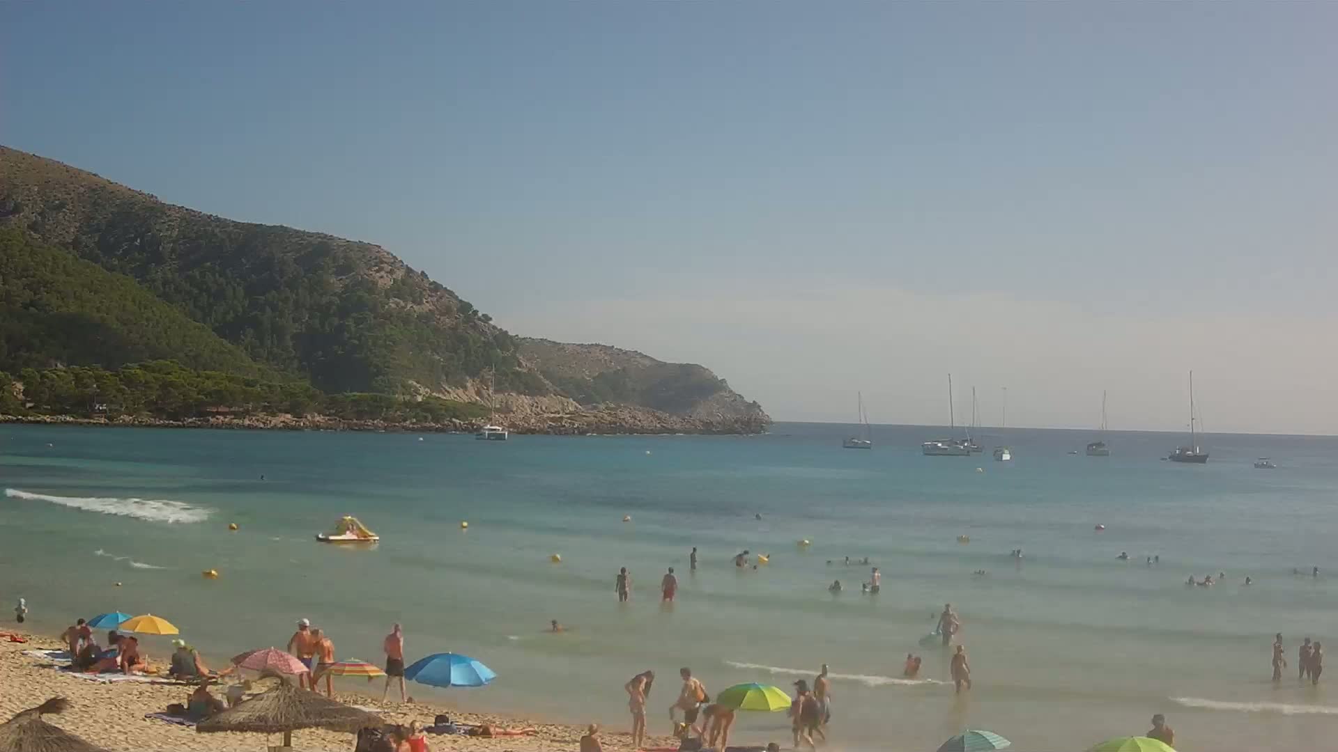 A sandy beach with people swimming and sunbathing under umbrellas, boats in the distance, and a clear, sunny day.
