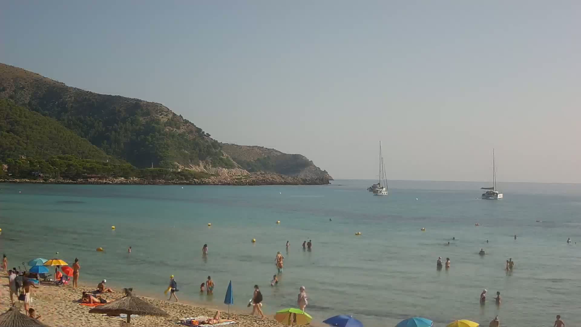 A sandy beach with people swimming and sunbathing under colorful umbrellas, clear turquoise water, and two sailboats in the distance on a sunny day.