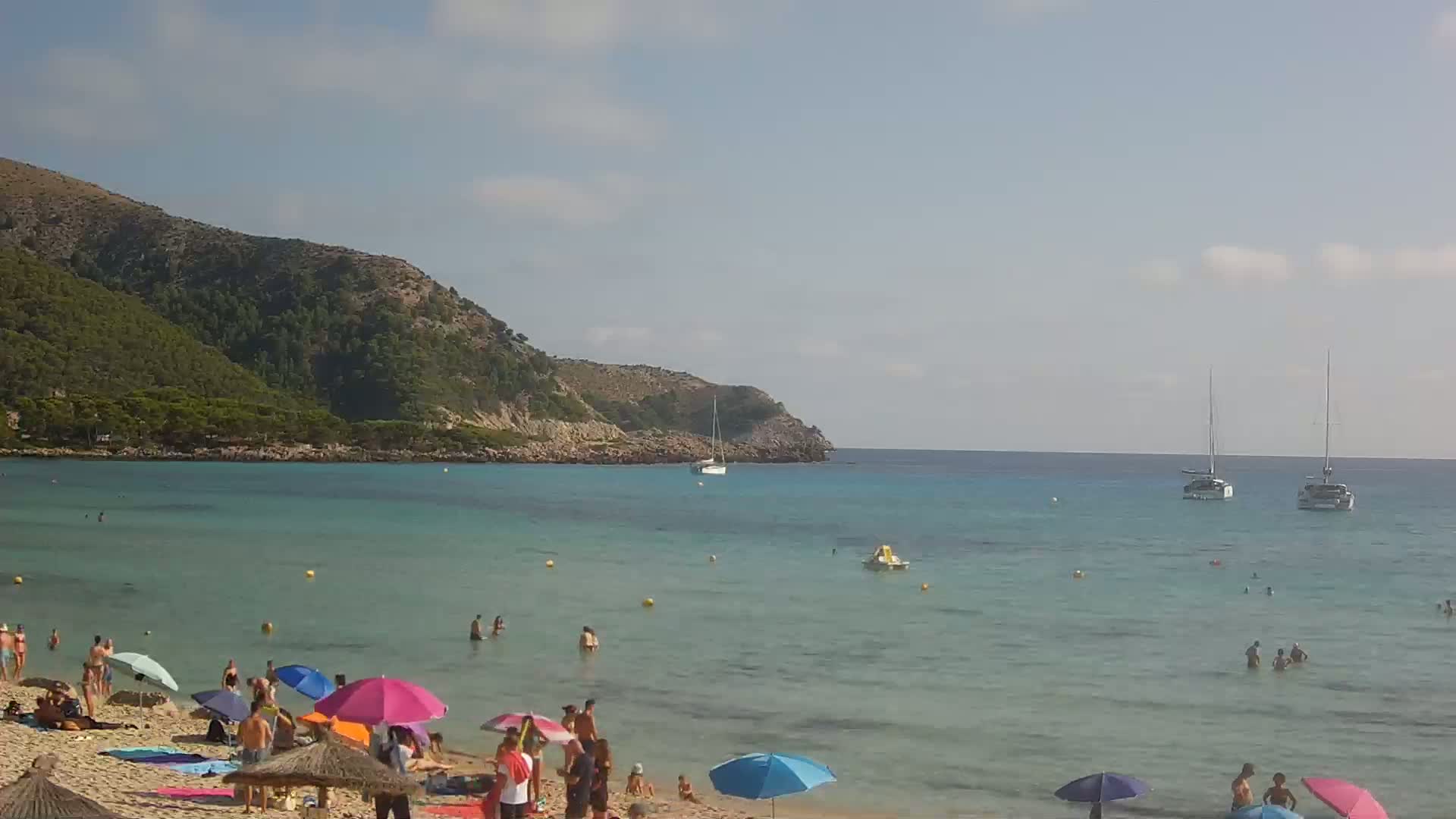 A sandy beach with colorful umbrellas and bathers is nestled below a green hill overlooking calm, clear turquoise water where several sailboats are anchored under a partly cloudy sky.