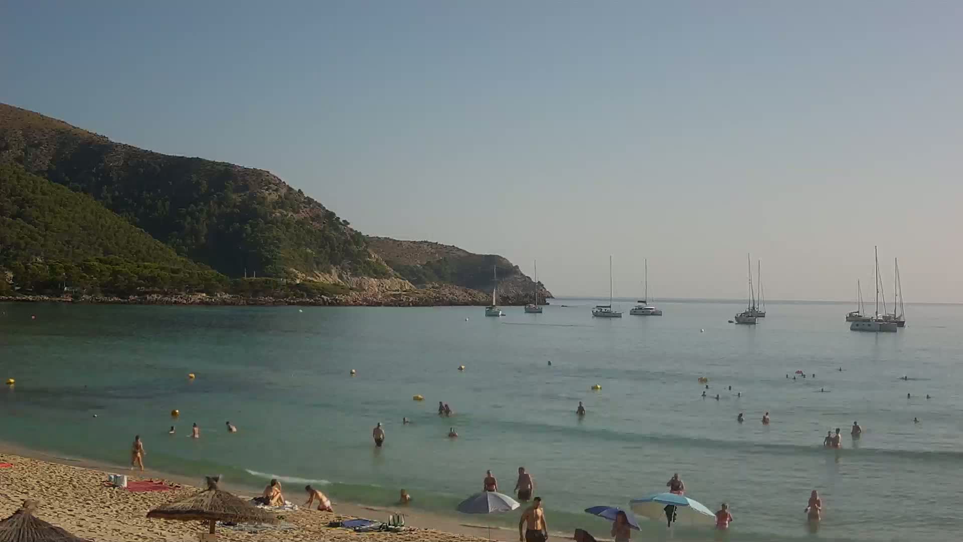 A sandy beach with people swimming and sunbathing under umbrellas is backed by a calm, clear sea with several sailboats anchored offshore and a verdant, rocky hill in the background under a clear, sunny sky.