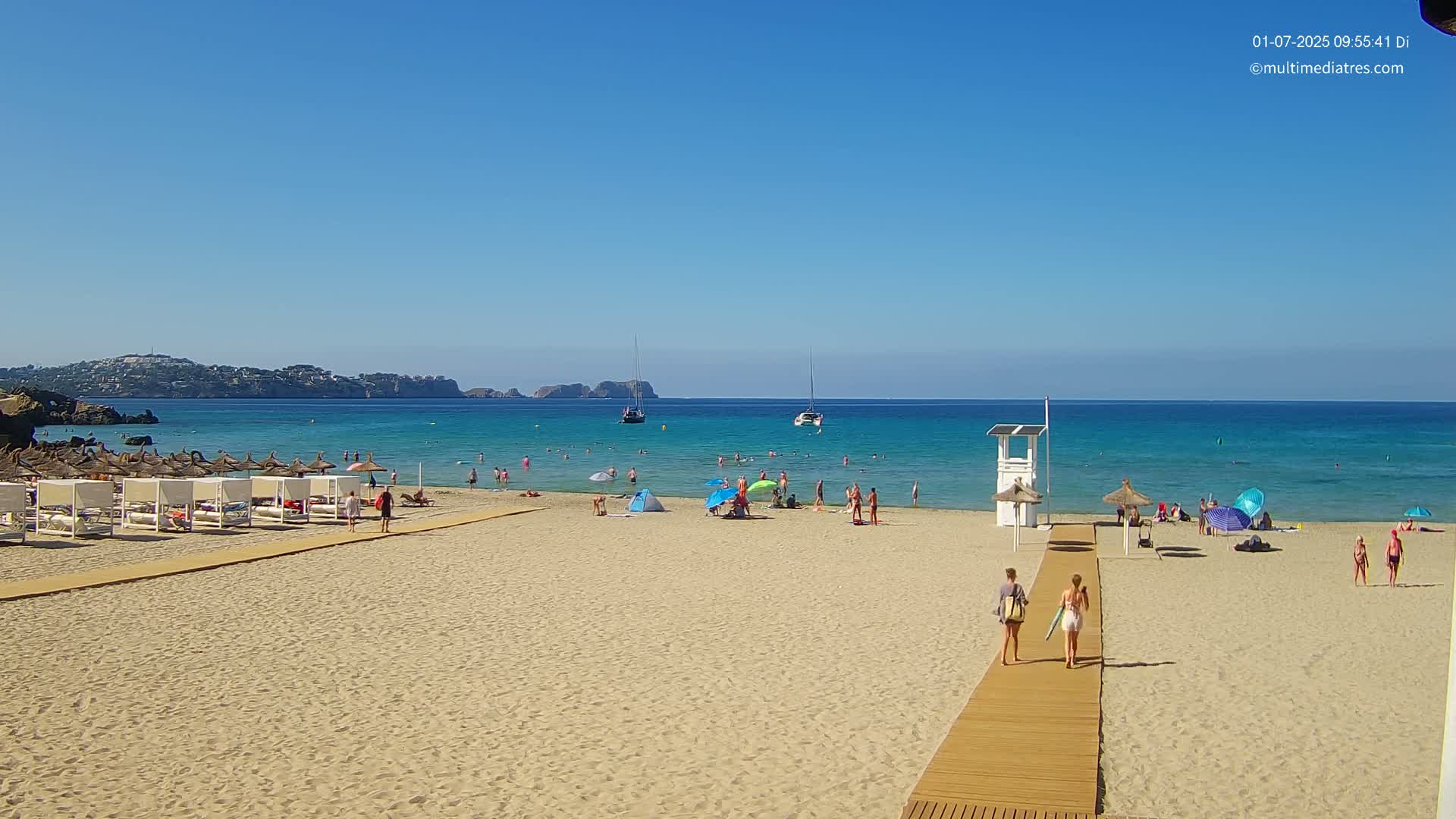 A sandy beach with people swimming and sunbathing, lined with beach umbrellas and lounge chairs, overlooks a calm, clear turquoise ocean under a bright, sunny sky.