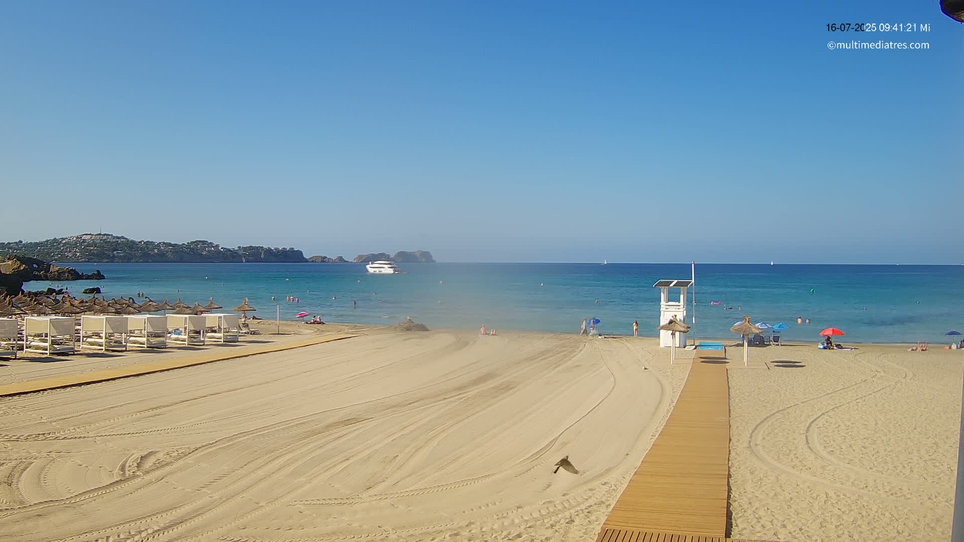 A sandy beach with a few people, beach umbrellas, and lounge chairs stretches towards a calm, clear blue sea under a bright sunny sky.