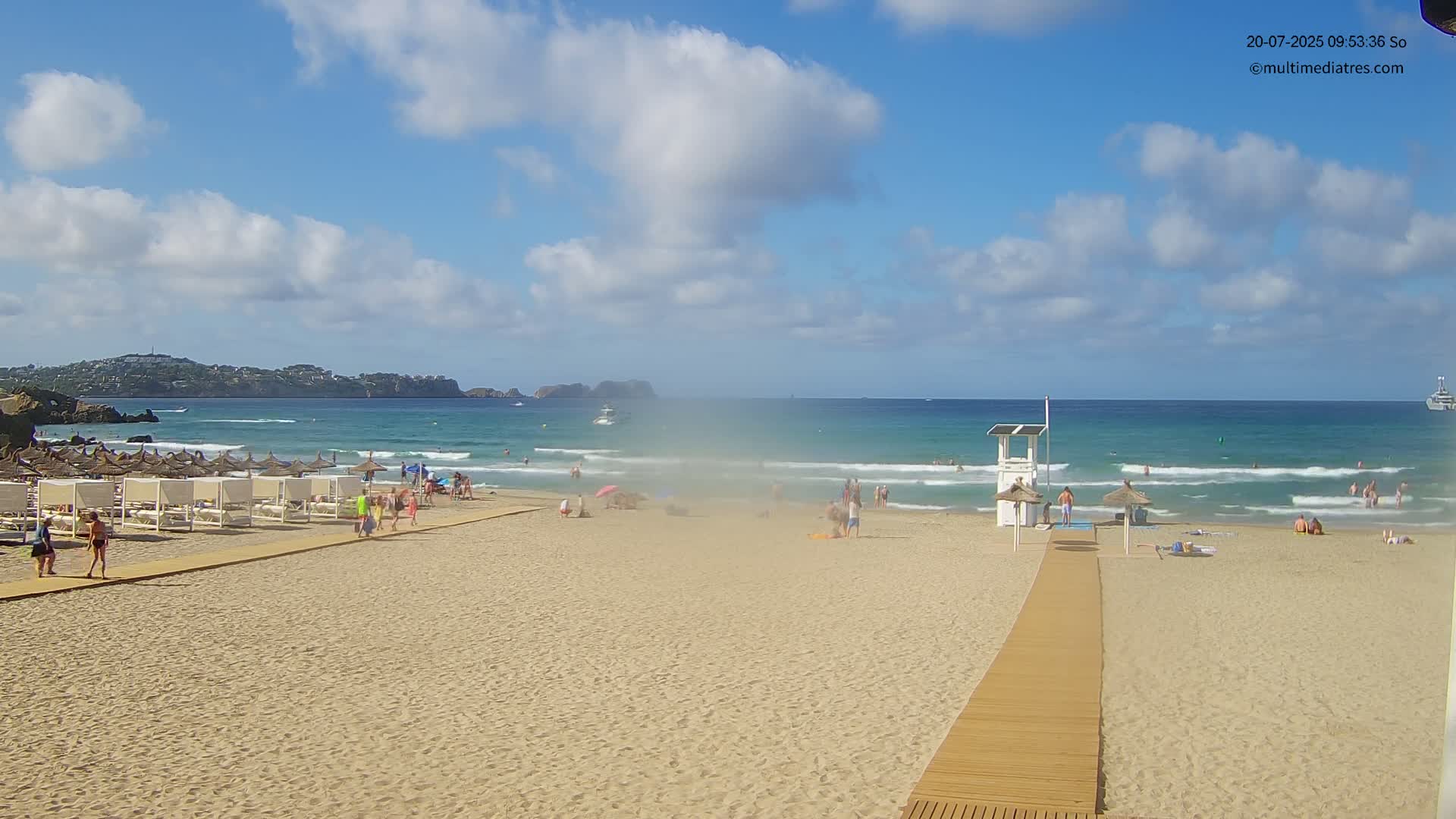 A sandy beach with a lifeguard stand, several people, and rows of beach umbrellas is seen under a partly cloudy sky, with a calm ocean in the background.