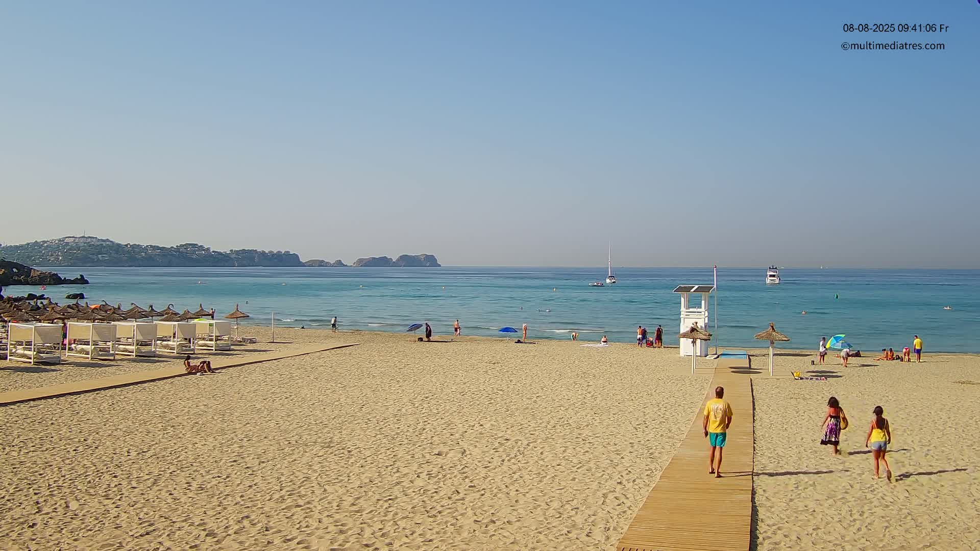 A sandy beach with several people and beach umbrellas under a clear, sunny sky, a lifeguard stand, and calm, blue water with boats in the distance.