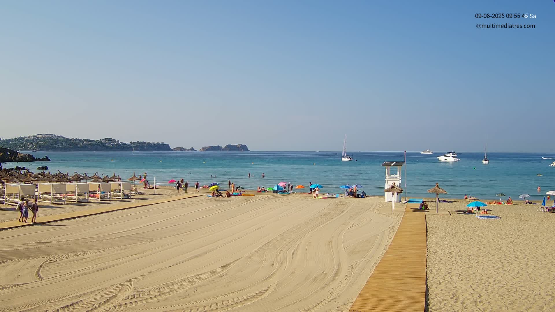 A sandy beach with several people, umbrellas, and beach cabanas stretches towards a calm, turquoise ocean under a clear blue sky.