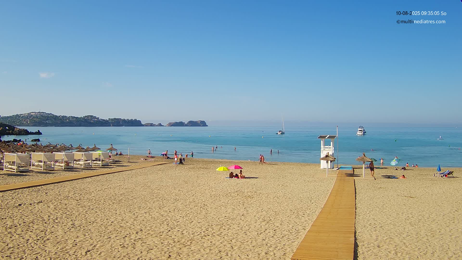 A sandy beach with people swimming and sunbathing under umbrellas, a lifeguard stand, and a wooden walkway leading to the calm, clear turquoise water under a clear blue sky.
