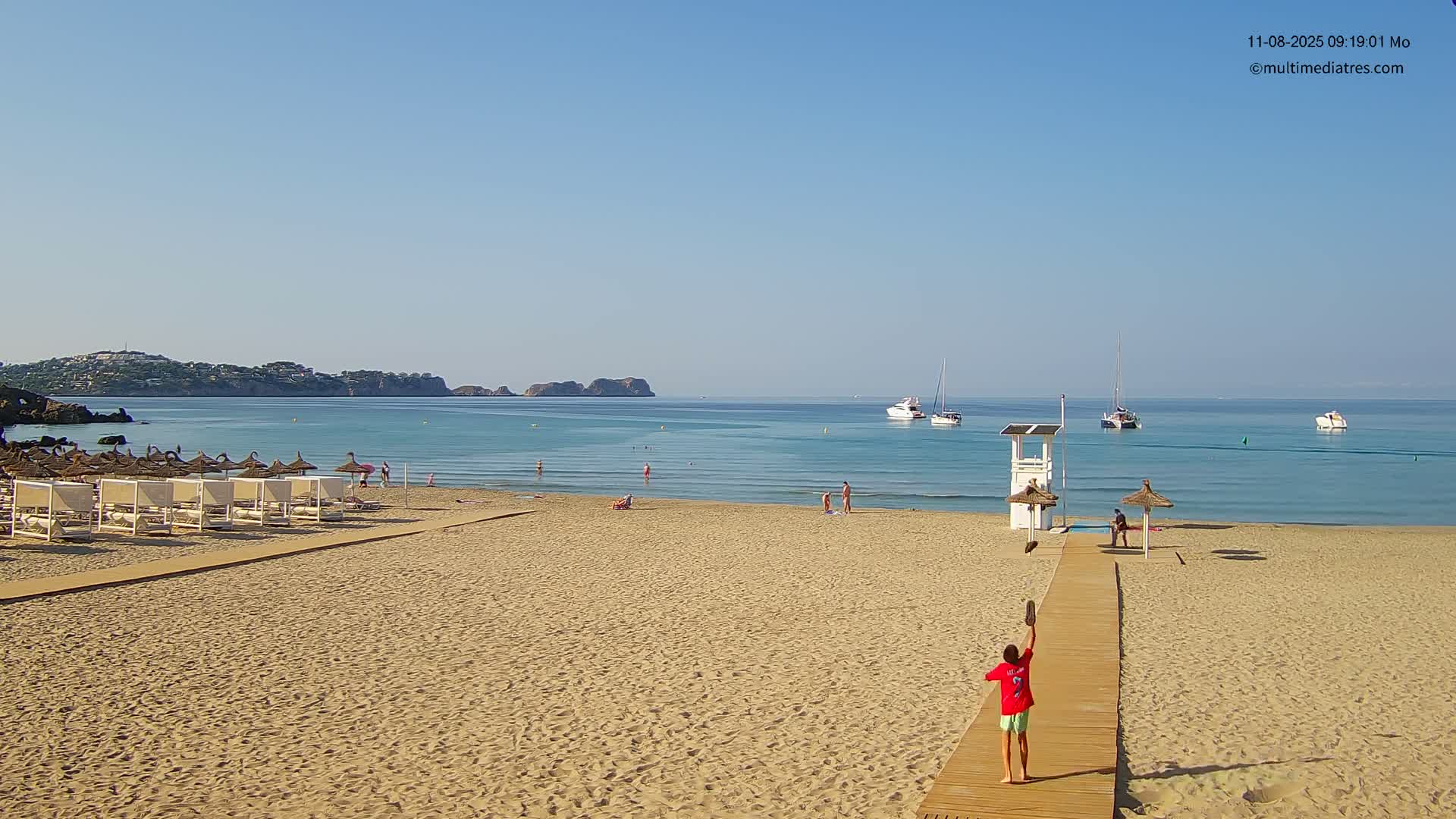 A sandy beach with several beach umbrellas and lounge chairs is seen on a sunny day, with a person standing on a wooden boardwalk tossing something in the air, boats in the calm water, and hills in the distance.