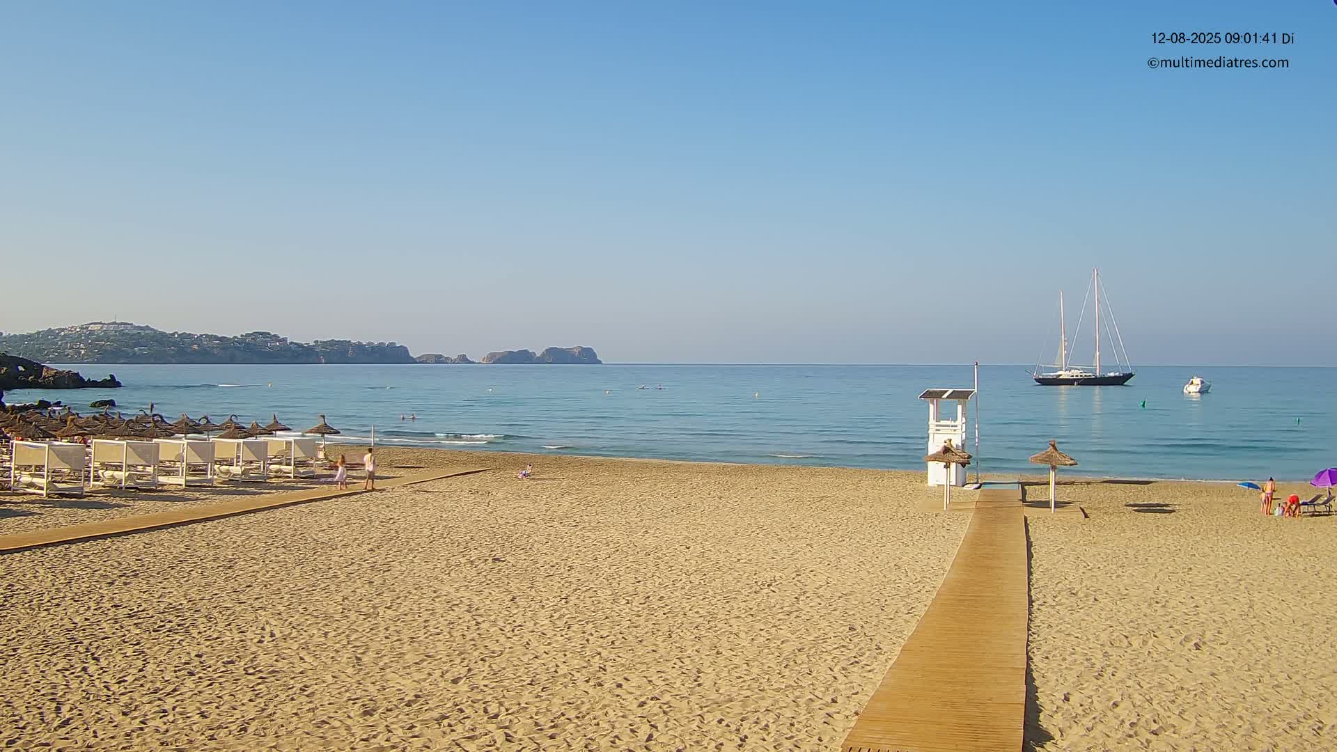 A sandy beach with a wooden walkway leading to a lifeguard station, several beach umbrellas and loungers, and a sailboat visible in the calm, clear water under a sunny sky.
