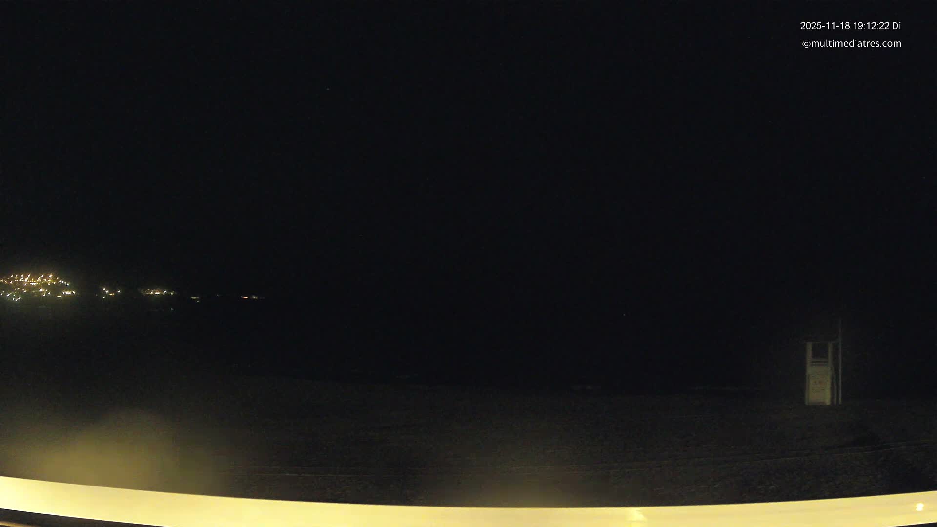 A sandy beach with a wooden walkway leading to a lifeguard station, several beach umbrellas and loungers, and a sailboat visible in the calm, clear water under a sunny sky.
