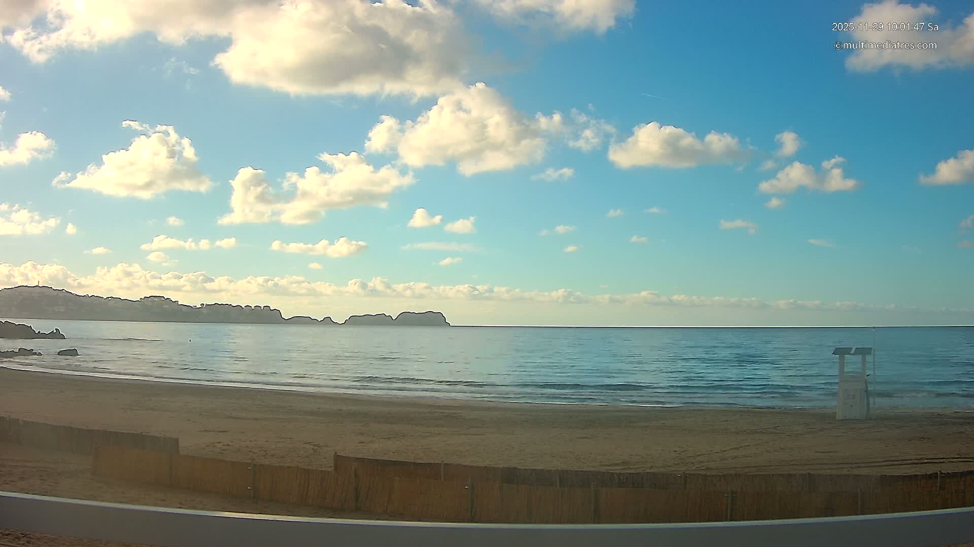 A scenic view depicts a sandy beach with a white lifeguard stand, calm blue ocean waters, and a distant hilly coastline, all under a bright blue sky with scattered white clouds on a partly sunny day.