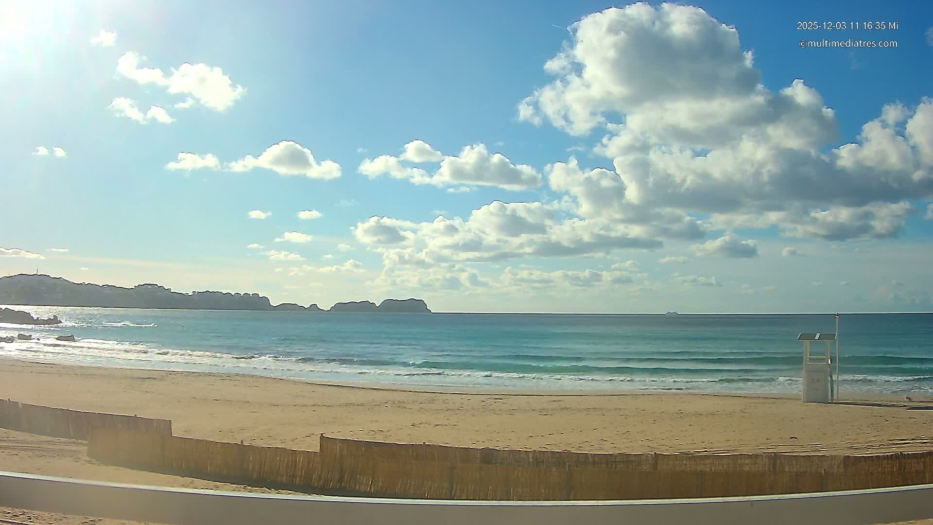A sunny beach scene reveals turquoise waves gently lapping a sandy shore with a lifeguard stand, a distant coastline, and a bright blue sky dotted with scattered white clouds.