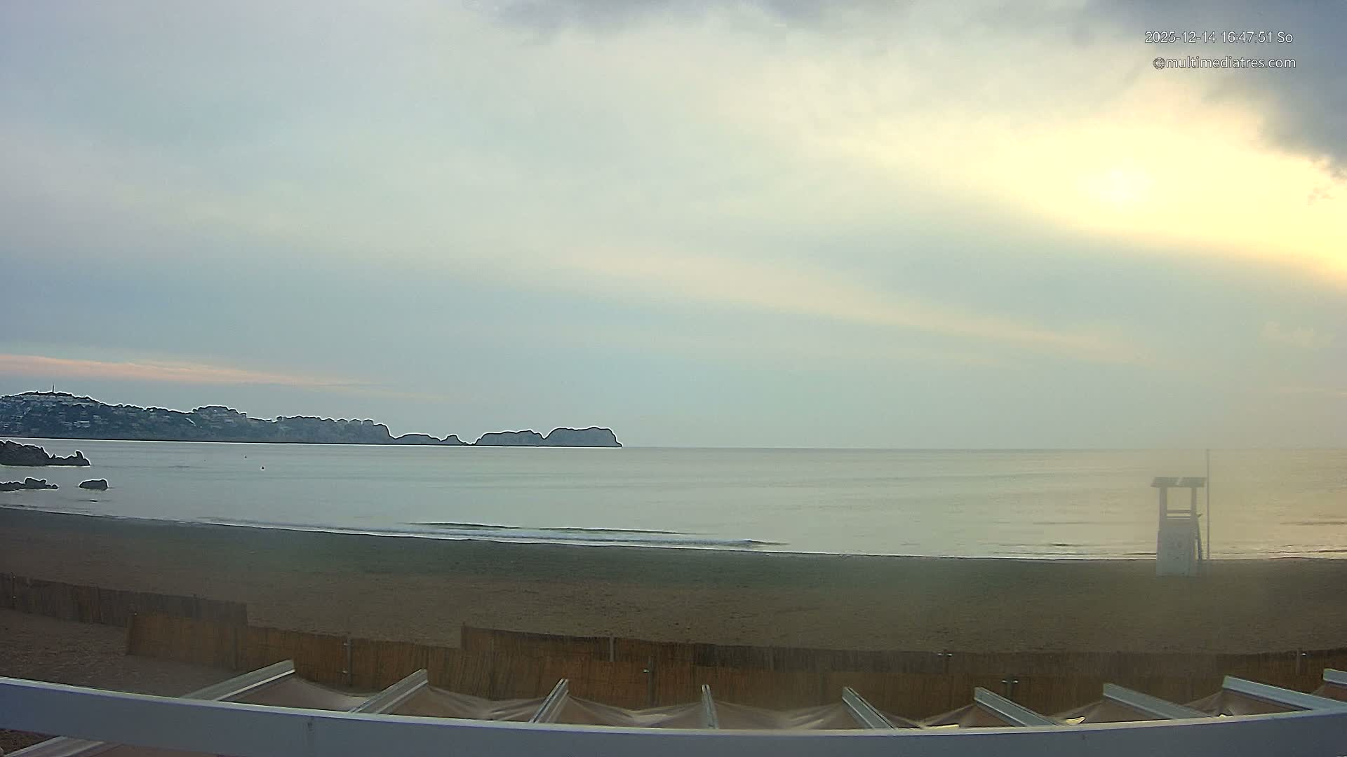 A sunny beach scene reveals turquoise waves gently lapping a sandy shore with a lifeguard stand, a distant coastline, and a bright blue sky dotted with scattered white clouds.