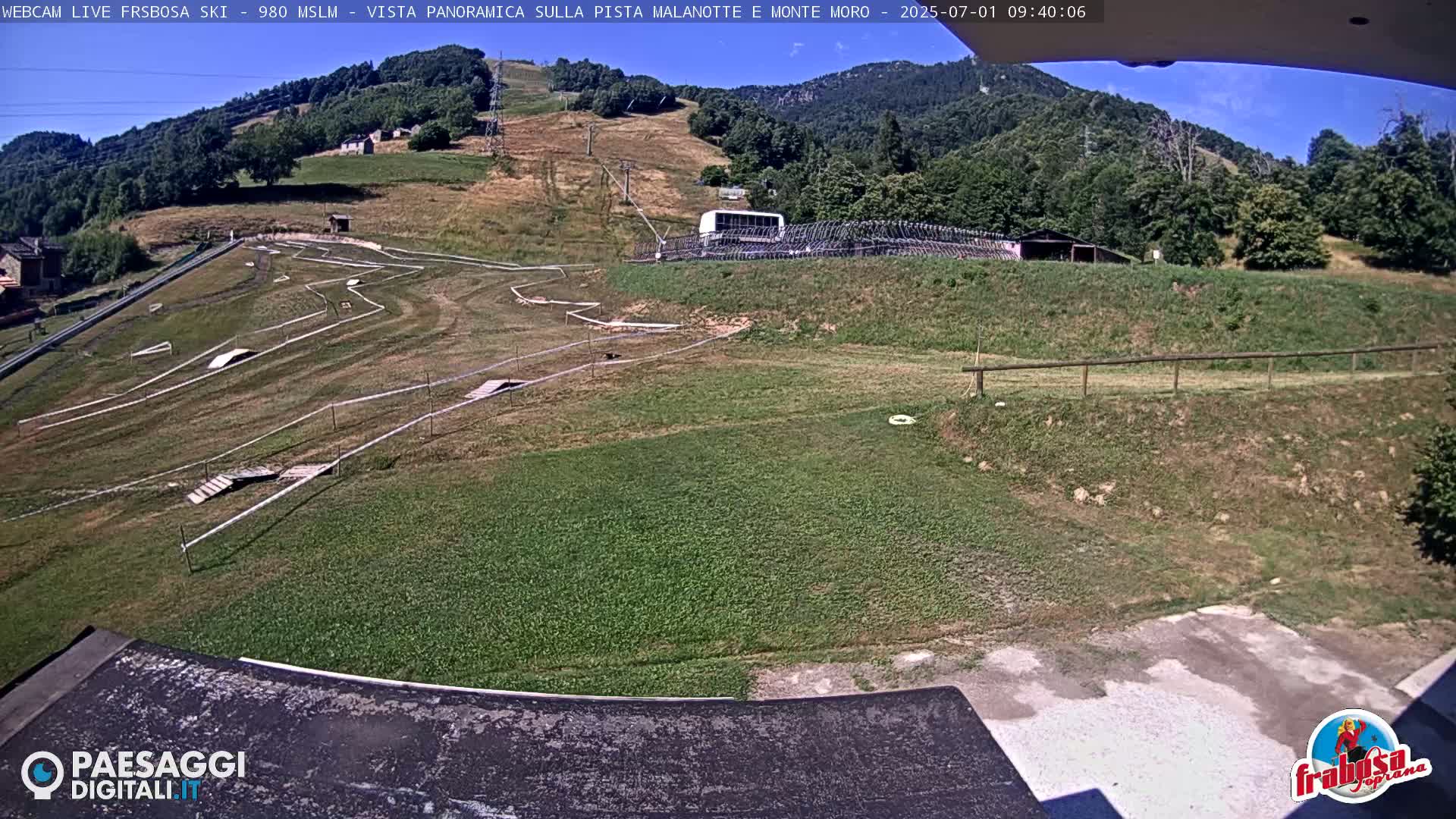 A panoramic view of a grassy hillside with an empty bike park and a bus parked uphill, under a mostly sunny sky.