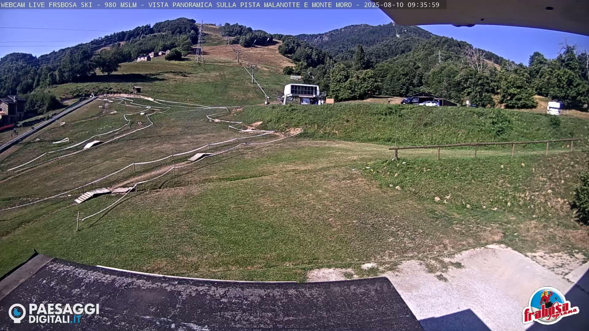 An outdoor view on a sunny day shows a grassy area with a bike track, a ski lift station, and a mountain in the background.