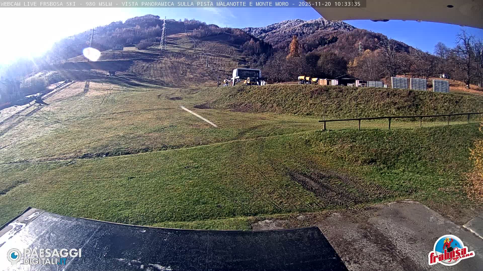 On a clear and sunny day, a mountain ski slope is visible, predominantly covered in green grass with tire tracks and a ski lift station, while the surrounding hillsides are dotted with trees showing late autumn colors, and the higher mountain peaks are dusted with snow.