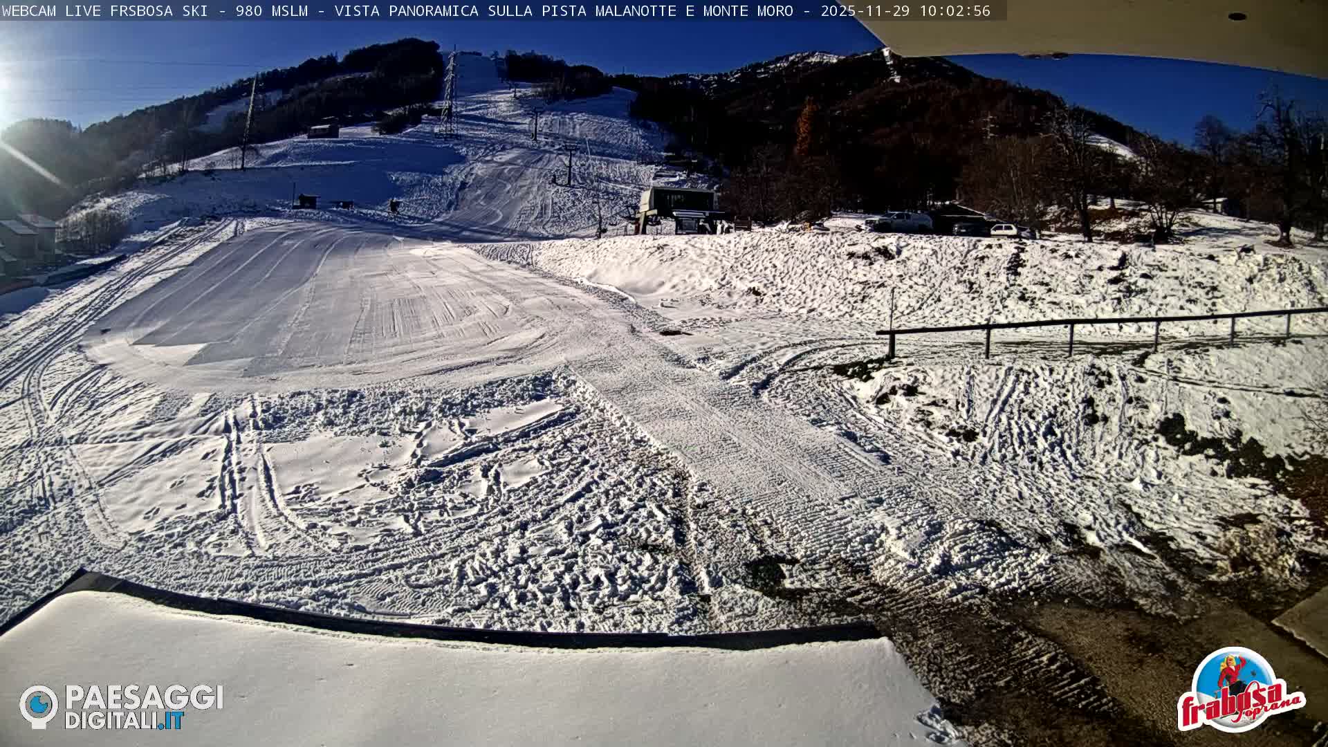 A sun-drenched winter scene at a ski resort features snow-covered slopes with visible ski tracks and lifts leading up a forested mountain under a clear blue sky.
