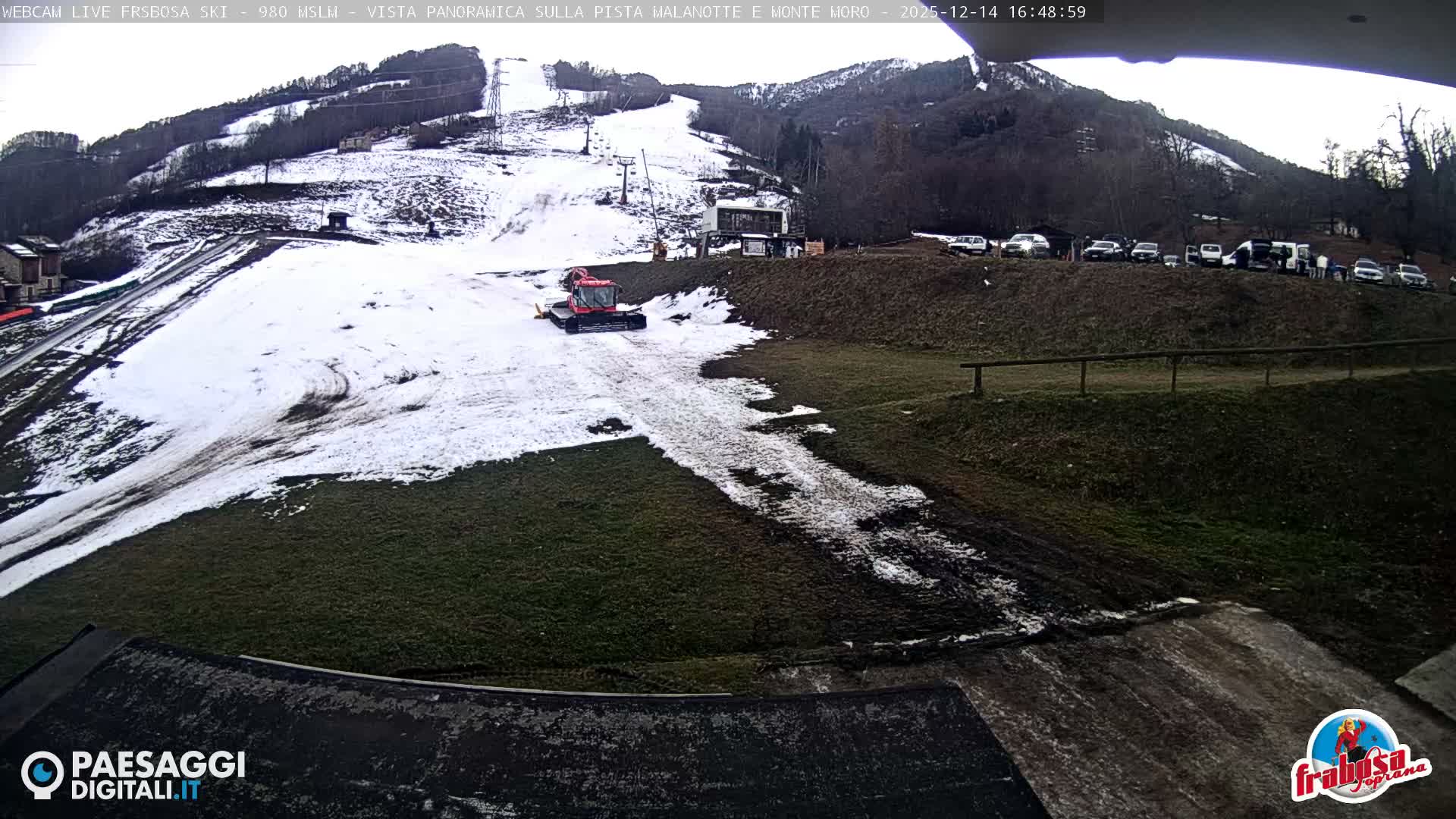 A sun-drenched winter scene at a ski resort features snow-covered slopes with visible ski tracks and lifts leading up a forested mountain under a clear blue sky.