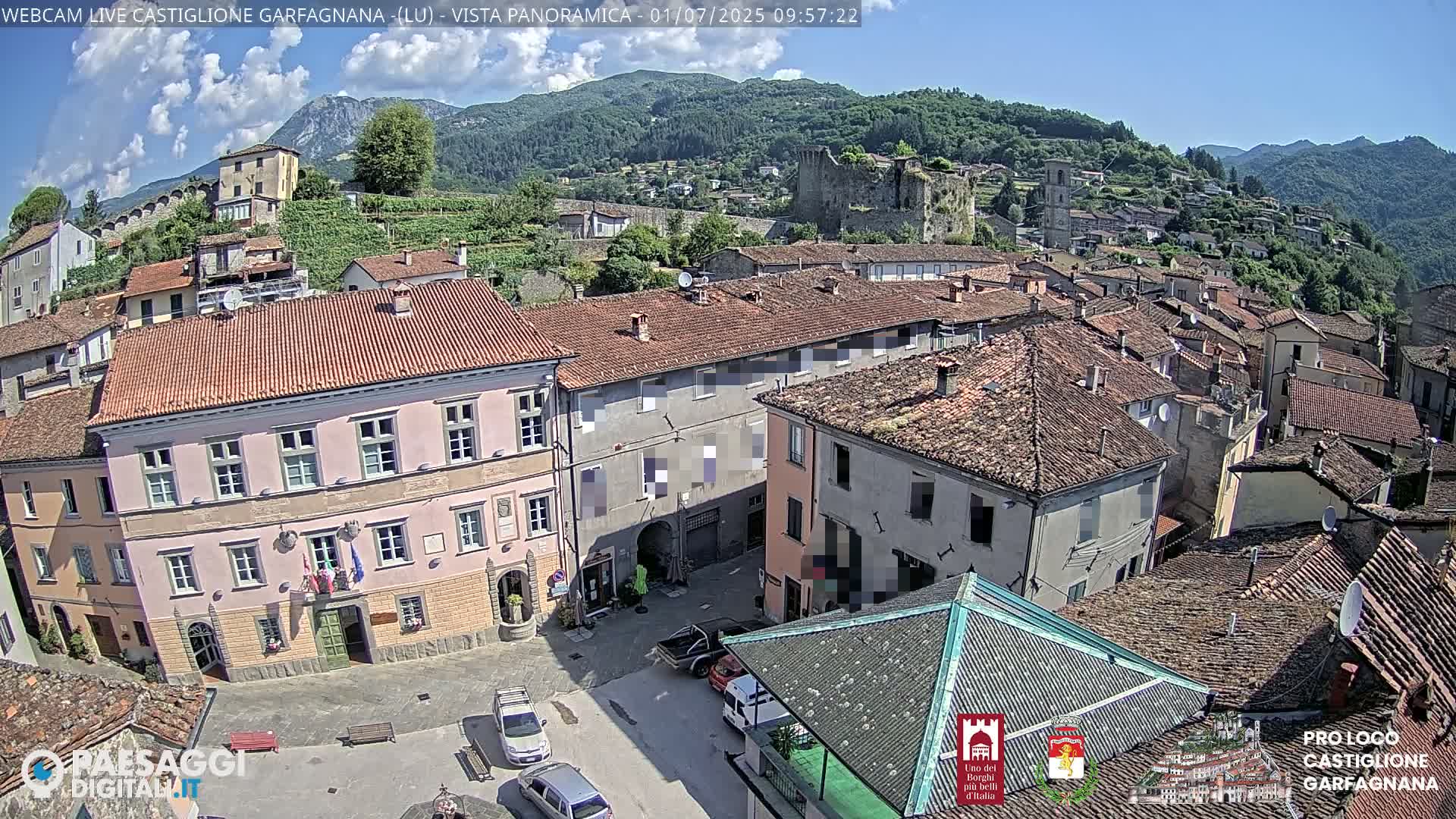 A picturesque town square with terracotta-roofed buildings is nestled in a valley, under a mostly sunny sky with a few clouds, and surrounded by hills and mountains.
