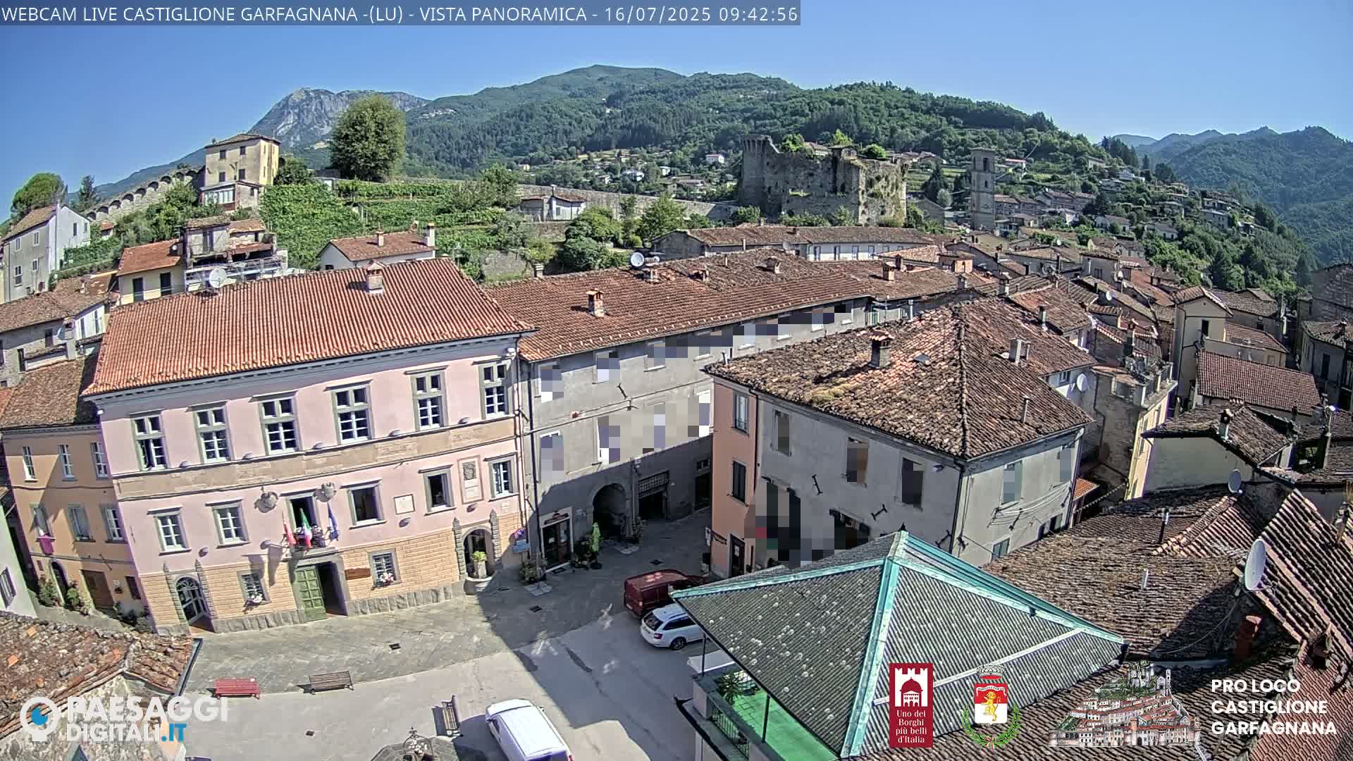 A sunny day reveals a town nestled in a valley, with red-tiled rooftops and a stone castle visible on a hillside.