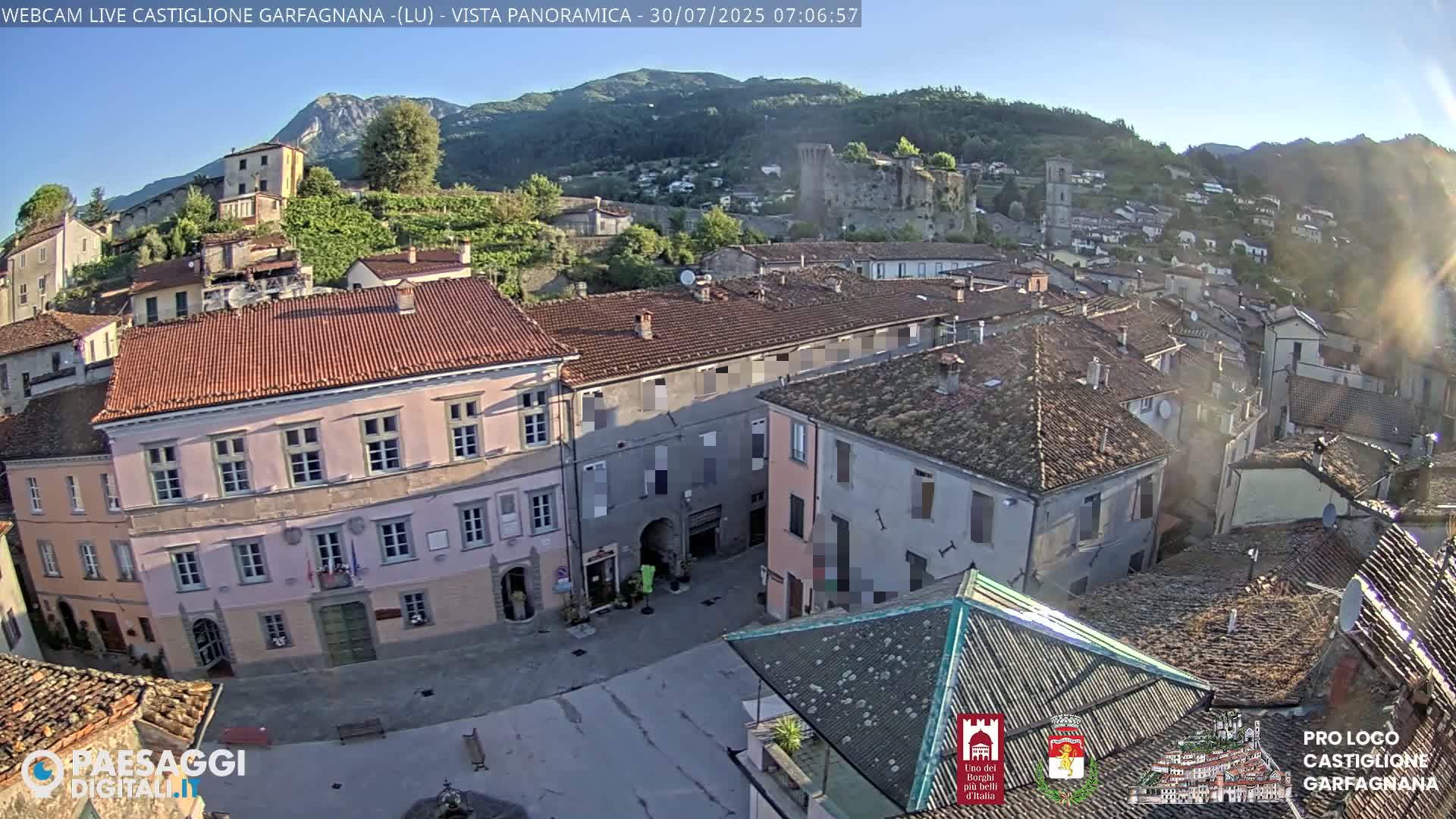 A mostly sunny day reveals a quaint town nestled in a valley, with a stone castle perched on a hillside in the background.