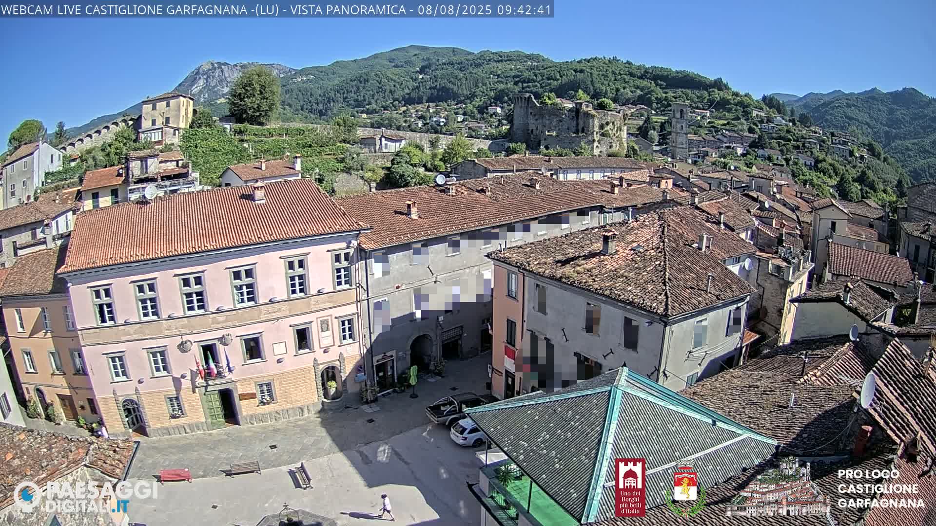 An aerial view shows a town square with buildings, some cars, and a person walking, under a clear, sunny sky, with hills and a castle visible in the background.