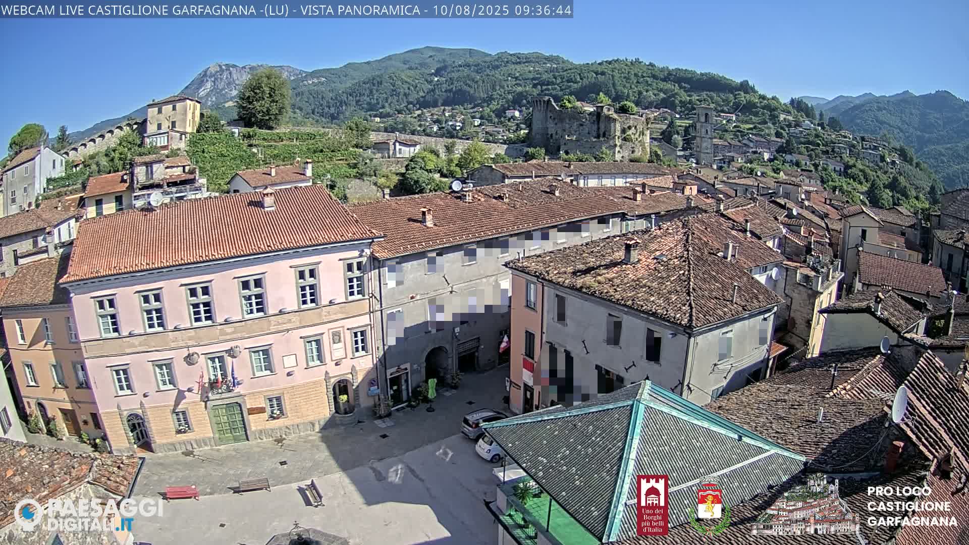 A sunny view shows a town nestled in a valley, with red-tiled roofs, a central plaza, and a hilltop castle in the background.