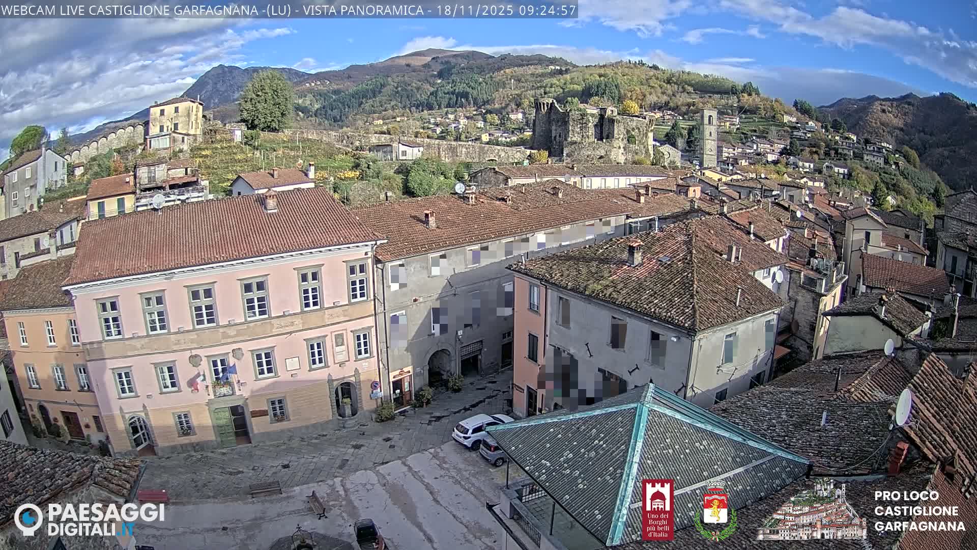 A panoramic view captures a historic hillside town with numerous red-tiled buildings, a prominent pink structure in the foreground, and ancient stone ruins on a distant forested mountain, all beneath a bright, partly cloudy blue sky.