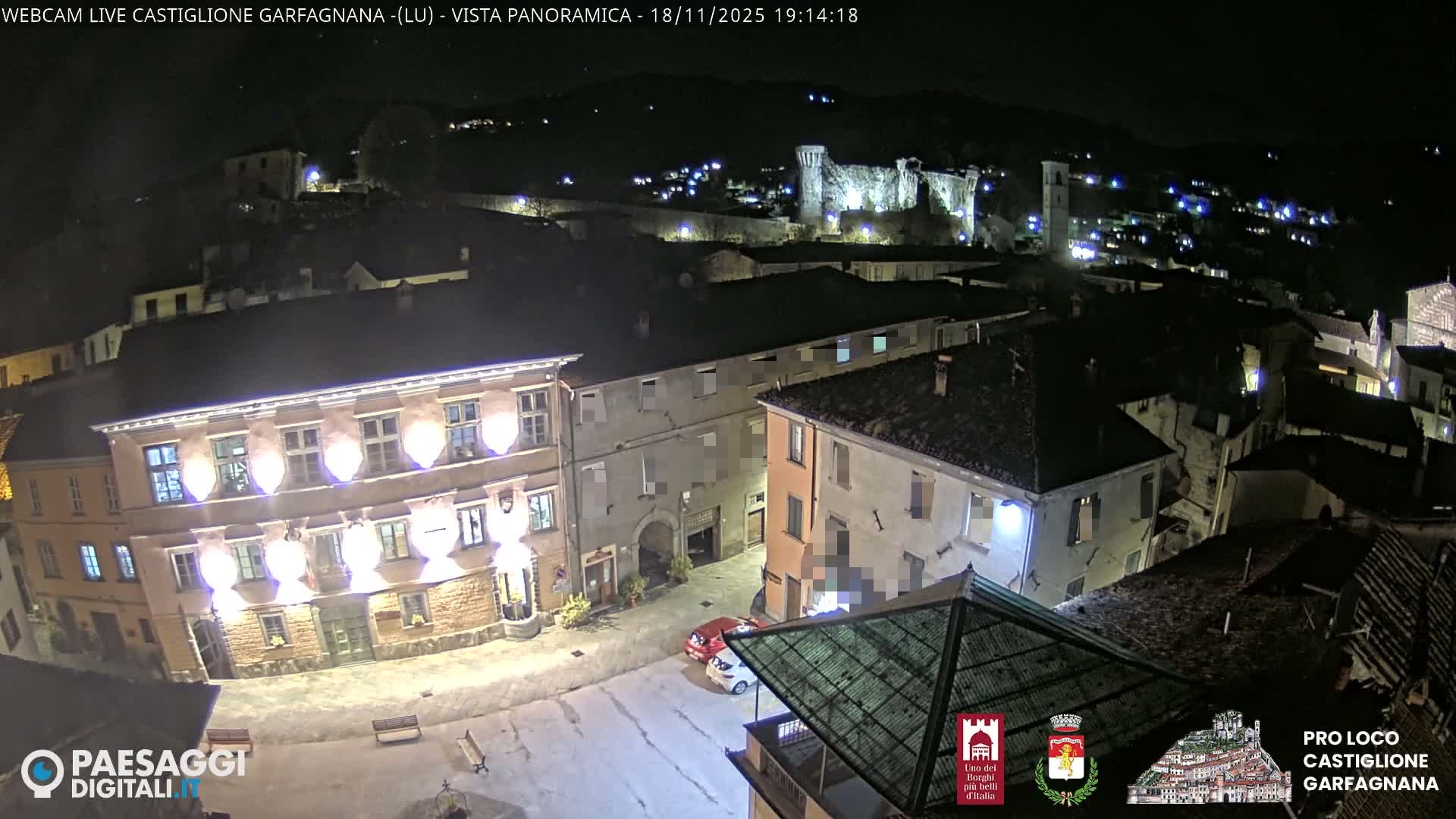 A panoramic view captures a historic hillside town with numerous red-tiled buildings, a prominent pink structure in the foreground, and ancient stone ruins on a distant forested mountain, all beneath a bright, partly cloudy blue sky.