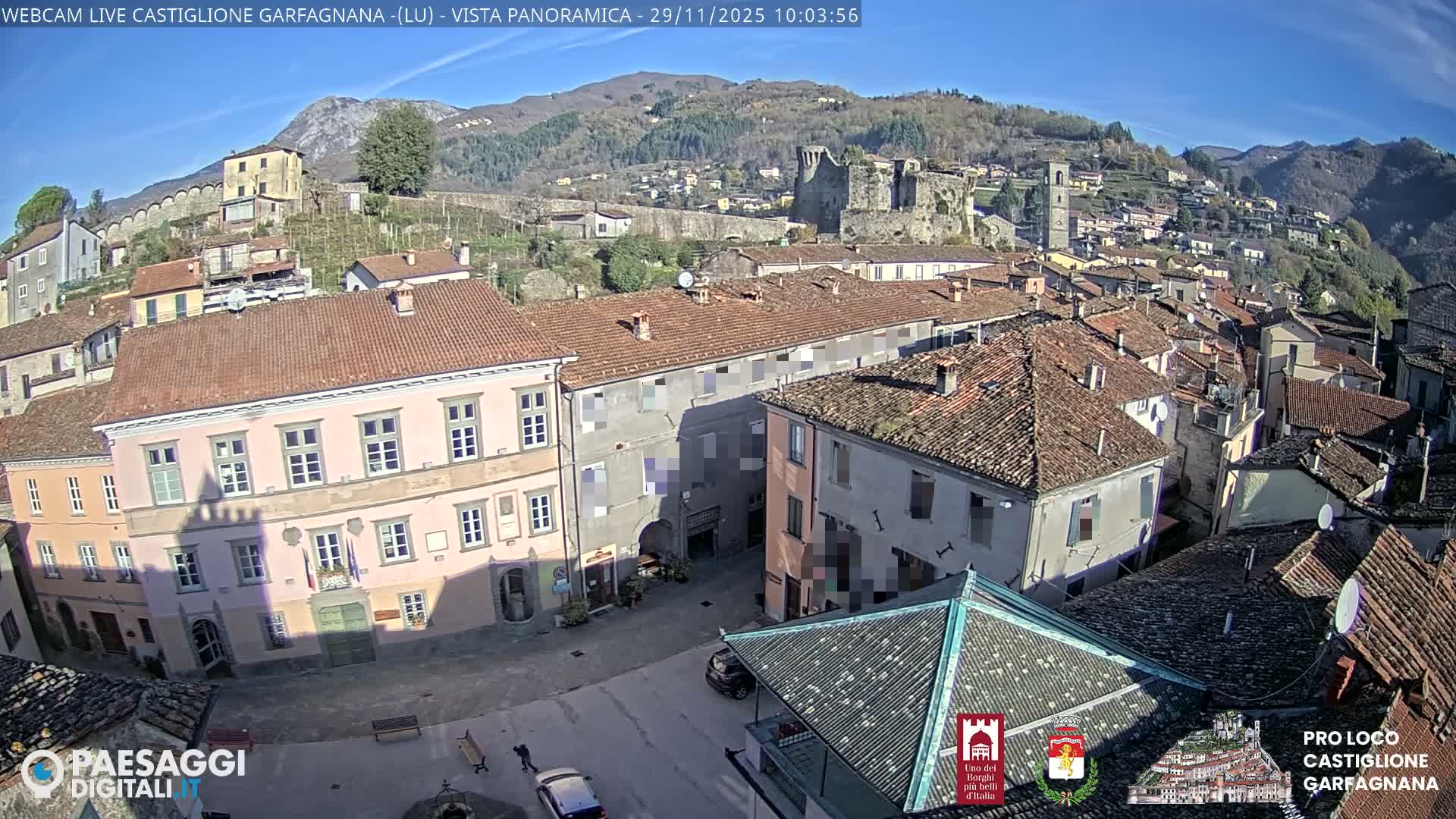 A panoramic view reveals a densely packed town with varied architecture and terracotta roofs, including a prominent pink building, nestled among green hills and mountains under a clear, sunny blue sky, with a castle ruin and a bell tower visible in the background.