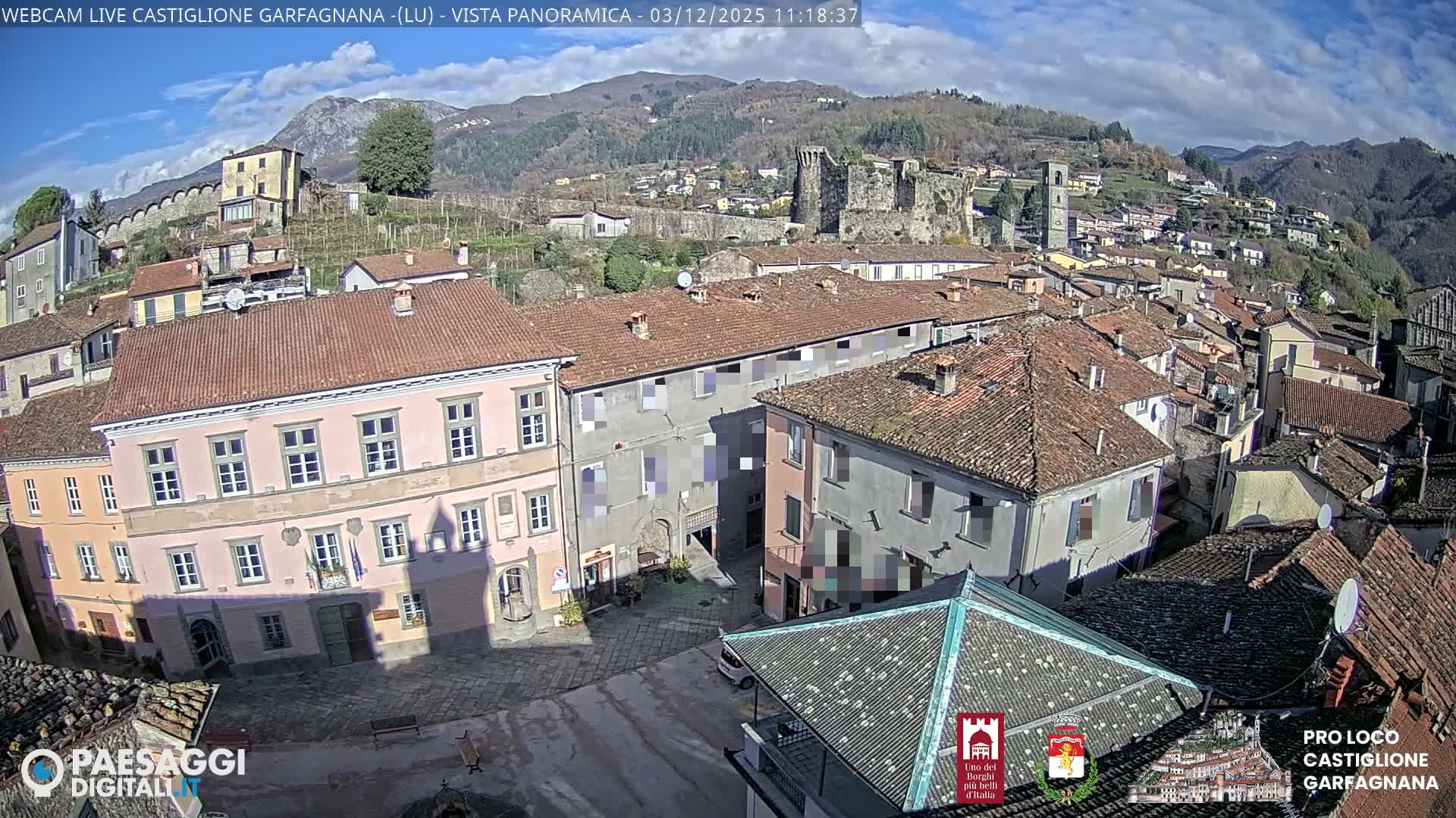 An elevated view captures a densely built town with numerous terracotta-roofed buildings, a central square, and a prominent castle ruin, nestled in a valley against a backdrop of forested mountains under a partly cloudy sky.