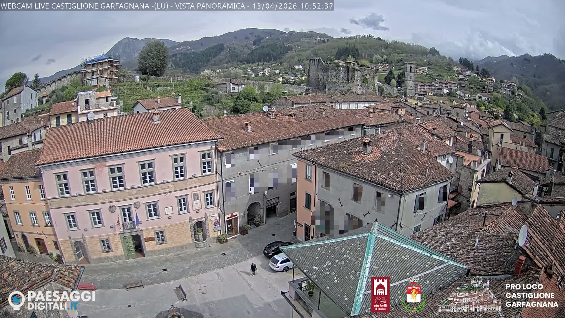 An elevated view captures a densely built town with numerous terracotta-roofed buildings, a central square, and a prominent castle ruin, nestled in a valley against a backdrop of forested mountains under a partly cloudy sky.